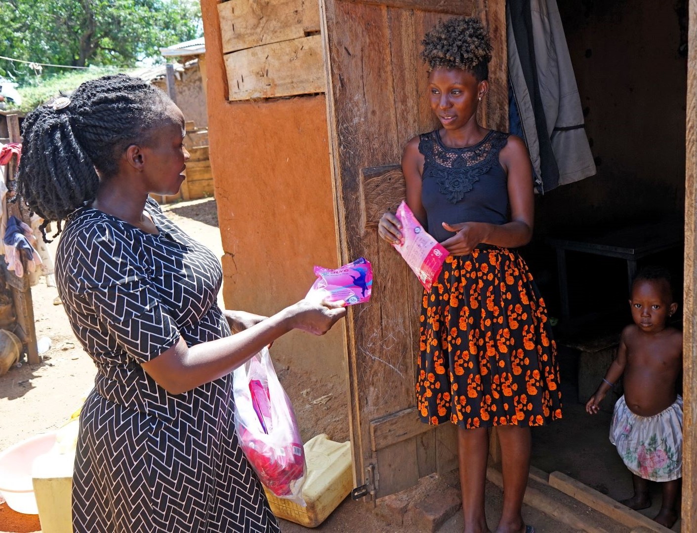 Zwei Frauen an einer offenen Haustür, die eine reicht der anderen ein pinkfarbenes Päckchen. Neben der Frau im Türrahmen steht noch ein Kleinkind.