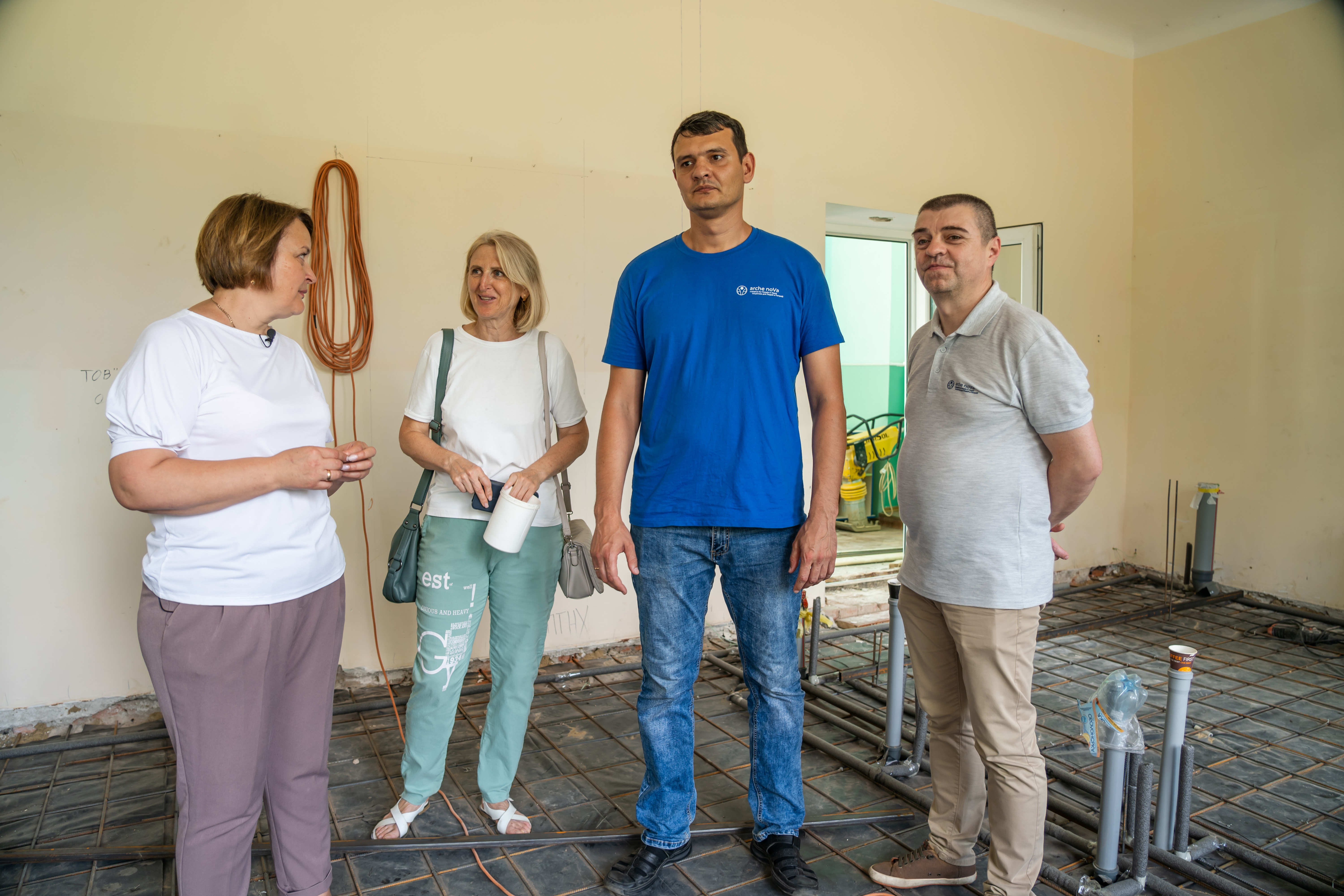 Das Foto zeigt vier Personen, zwei Frauen und zwei Männer, in einem Raum, in dem Baumaßnahmen am Boden durchgeführt werden. Aus dem Boden ragen an einigen Stellen Rohre.