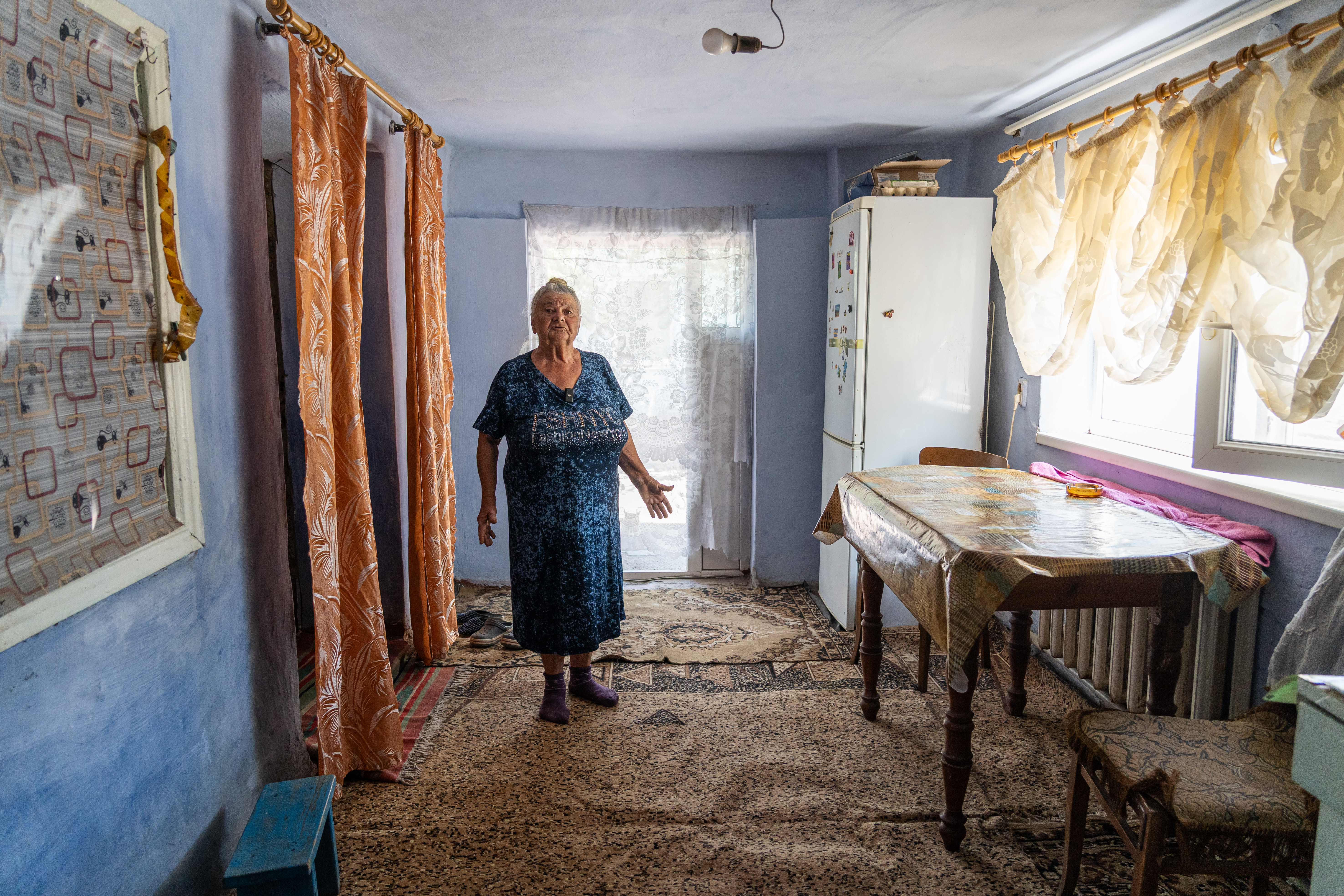 Das Foto zeigt eine kleine Frau im hohen Alter in einem Raum stehend. Am Boden liegt ein Teppich, rechts stehen ein Kühlschrank, ein Tisch und ein Stuhl.
