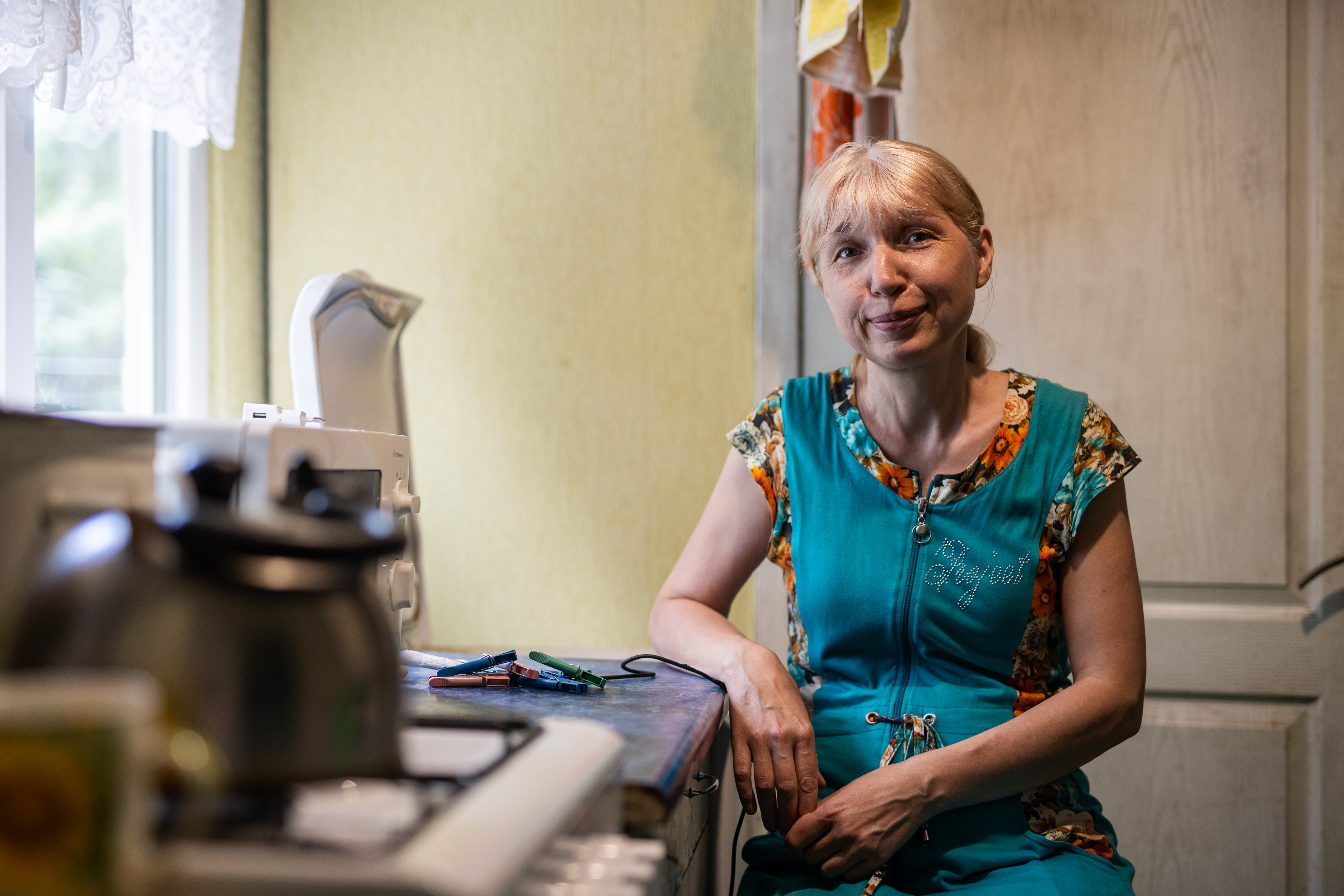 Eine Frau mittleren Alters sitzt neben einem Küchenherd. Sie trägt ein blaues Kleid, darunter eine kurzärmelige Bluse mit Blumen. Ihr weißblondes Haar trägt sie zusammengebunden.