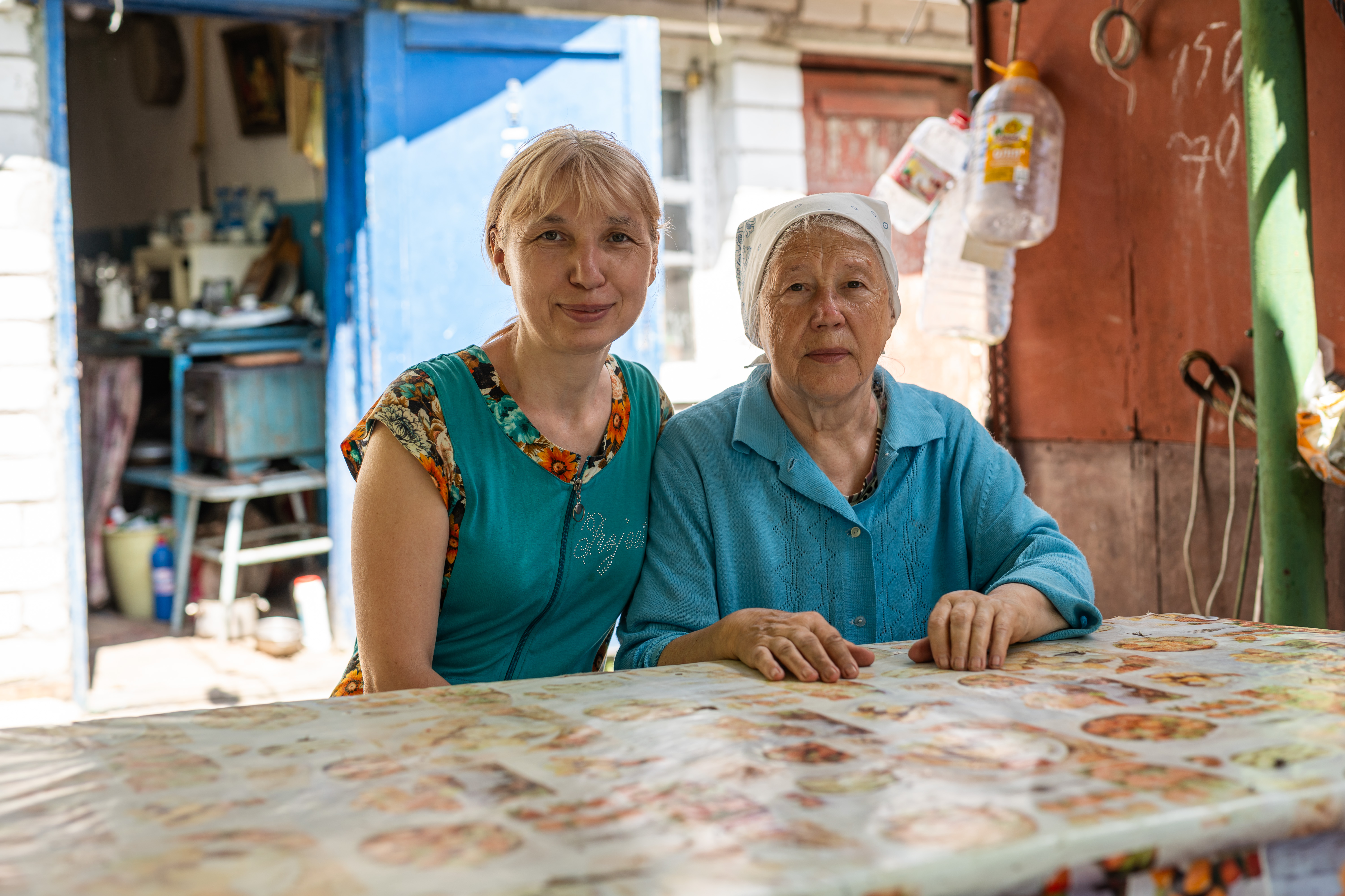 Das Bild zeigt zwei Frauen, eine im mittleren, eine im höheren Alter. Sie sitzen draußen an einem Tisch, im Hintergrund scheint die Sonne. Sie wirken einander zugewandt und sitzen nah beieinander.