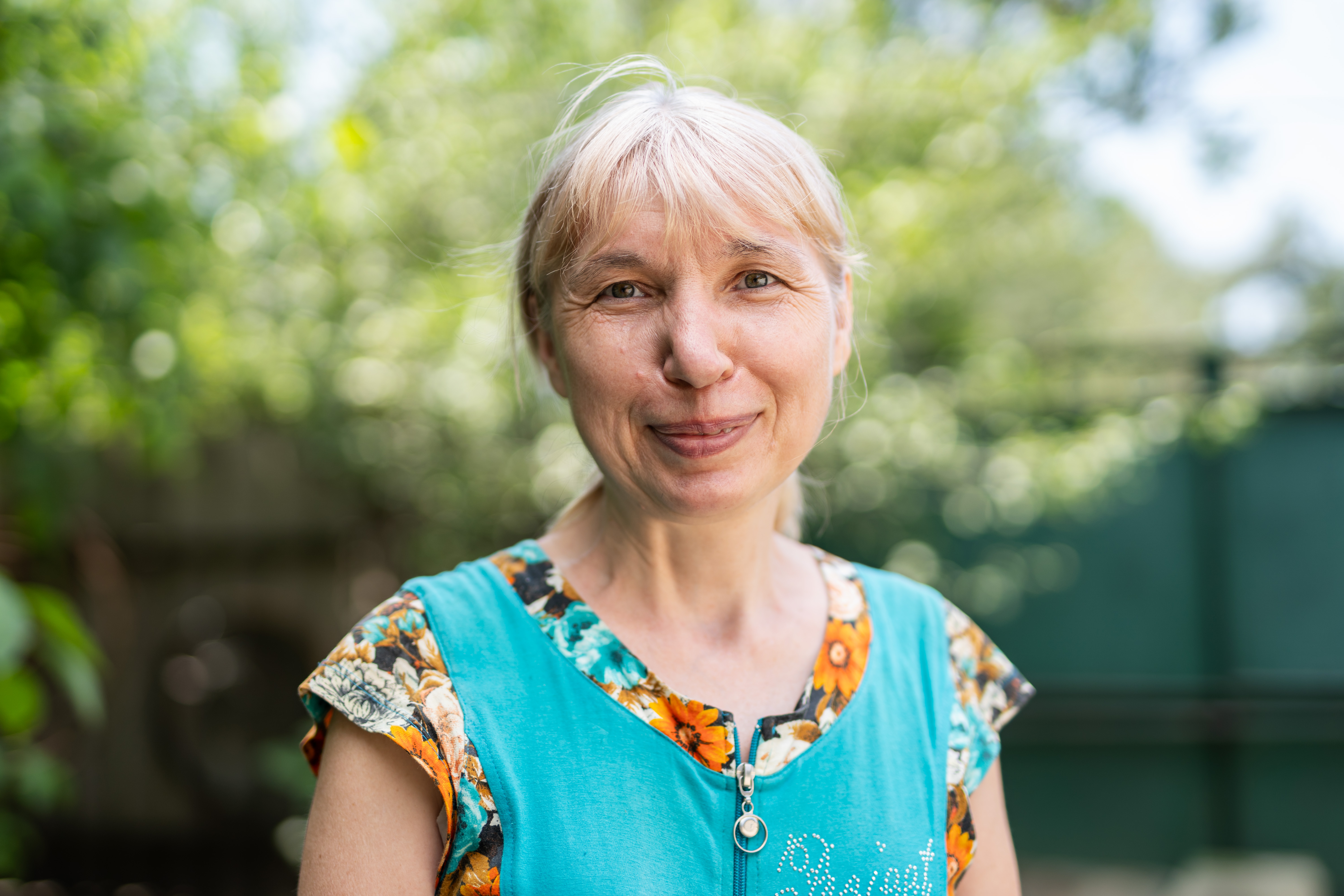 Das Foto zeigt eine Frau mittleren Alters mit hellen Haaren und bunter Kleidung im Portrait. Sie lächelt in die Kamera, im Hintergrund sind unscharf Bäume zu sehen.