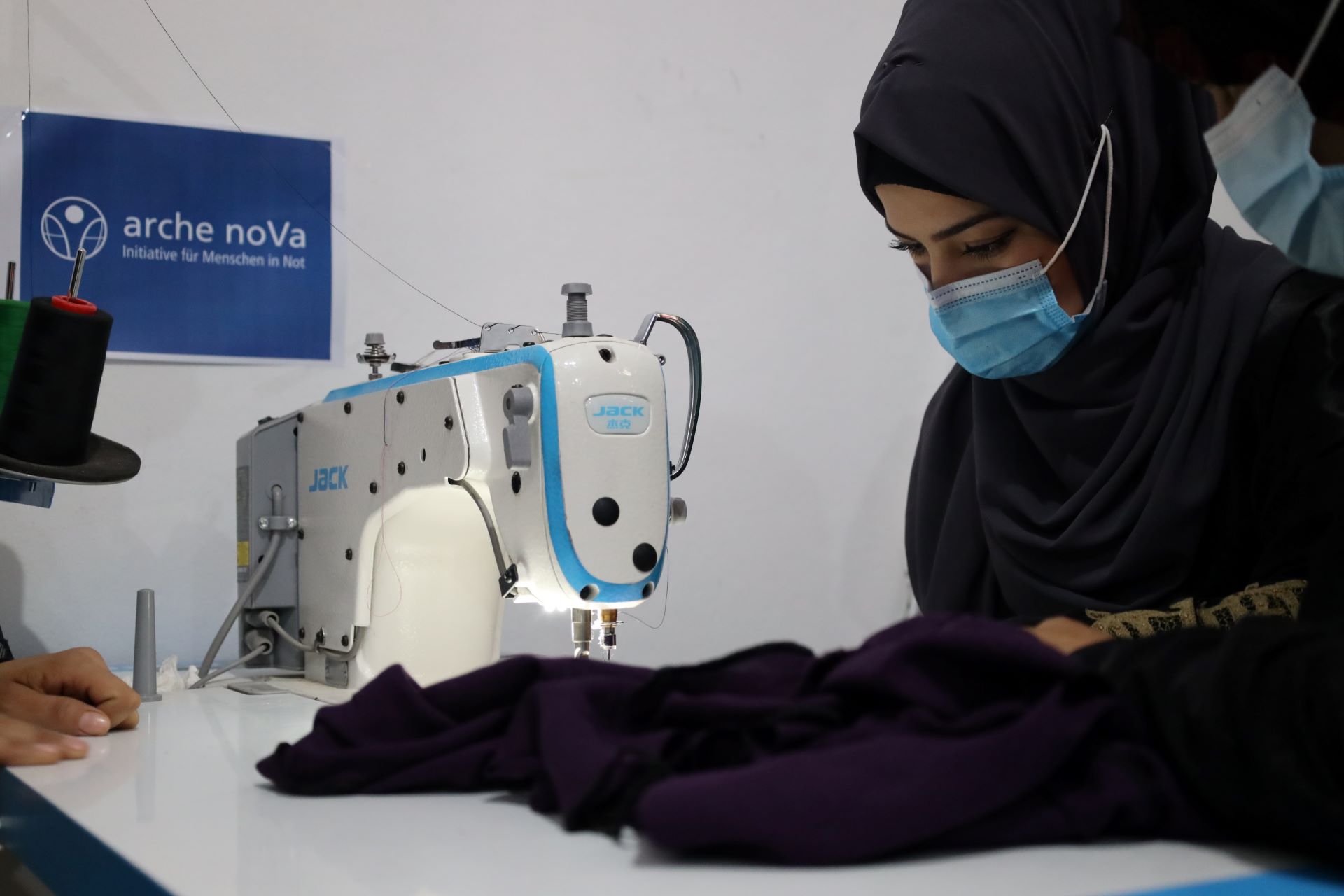 Das Bild zeigt eine junge Frau, die eine Nähmaschine bedient. Im Hintergrund ist das Logo von arche noVa zu sehen.
