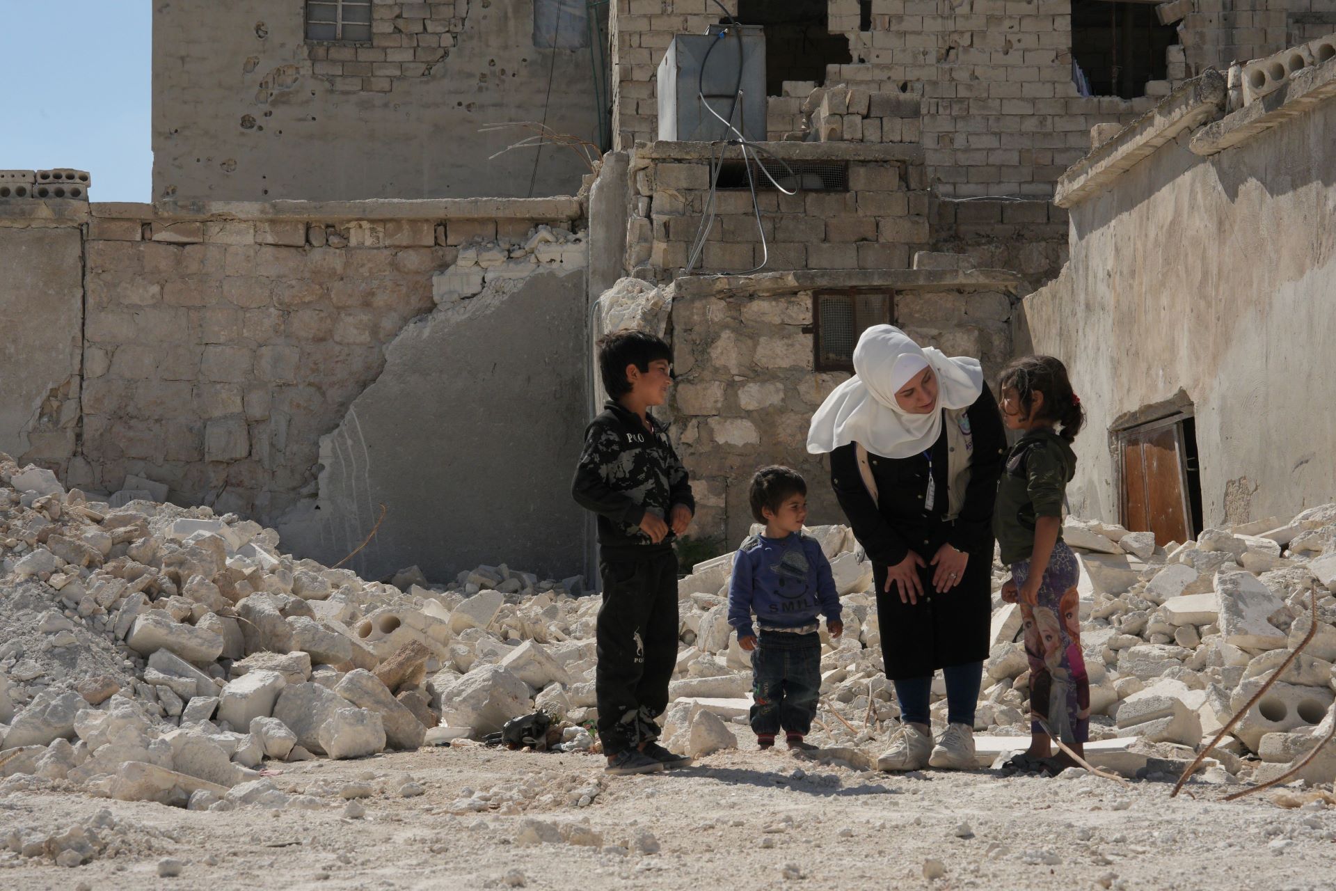 Das Bild zeigt die Bonyan-Helferin Sana umgeben von drei Kindern unterschiedlichen Alters. Sie stehen inmitten von Trümmern, Sana beugt sich zu einem der Kinder hinunter.