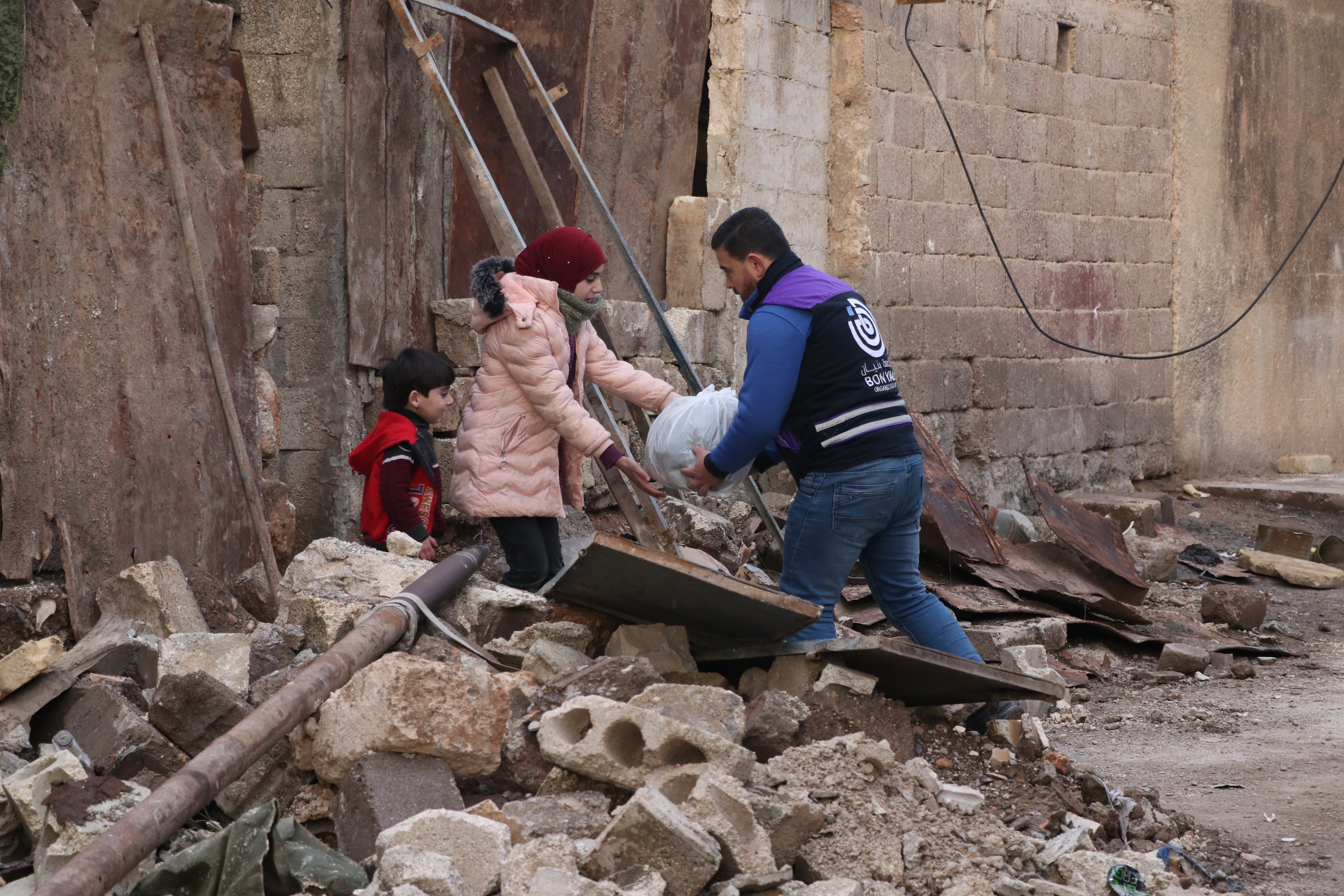 Mann übergibt eine Tüte mit Hilfsgütern an eine Frau. Daneben steht ein kleines Kind. Im Vordergrund sind Trümmer zu sehen.