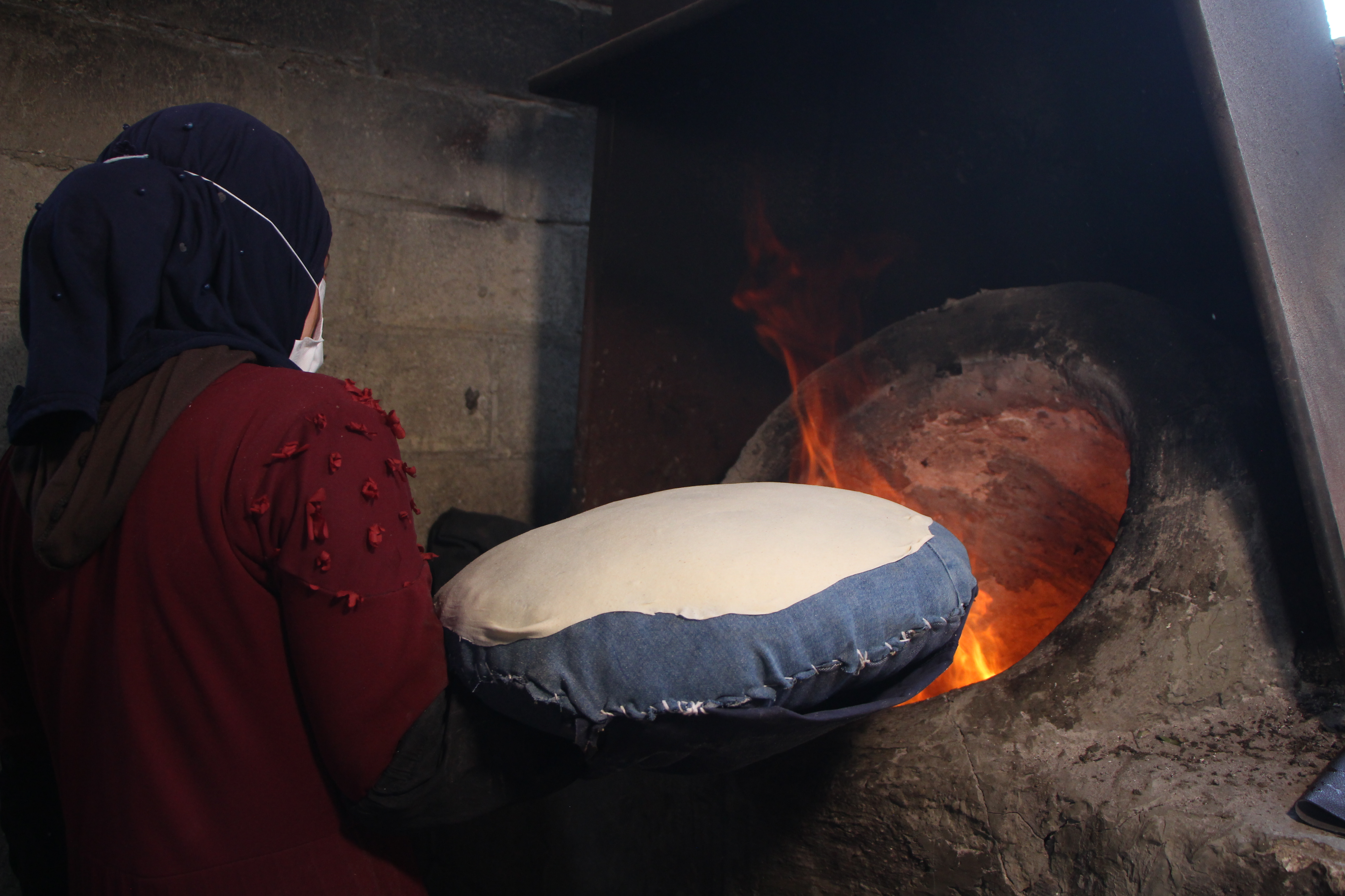 Eine Frau hält ein Fladenbrot auf einem Kissen ausgebreitet vor einem offen Backofenfeuer