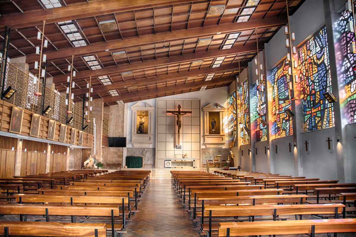 Innenansicht eines modernen Kirchengebäudes mit Schrägdach und bunten Fenstern rechts, durch die Licht einfällt.