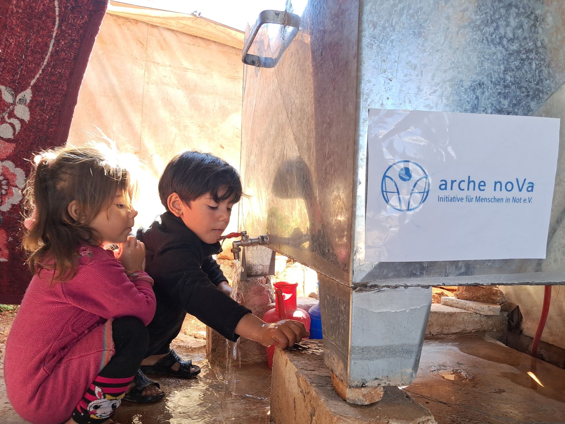 Das Bild zeigt zwei Kinder neben einem Wassertank aus Metall, die sich die Hände waschen. Auf dem Tank ist das Logo von arche noVa zu sehen.