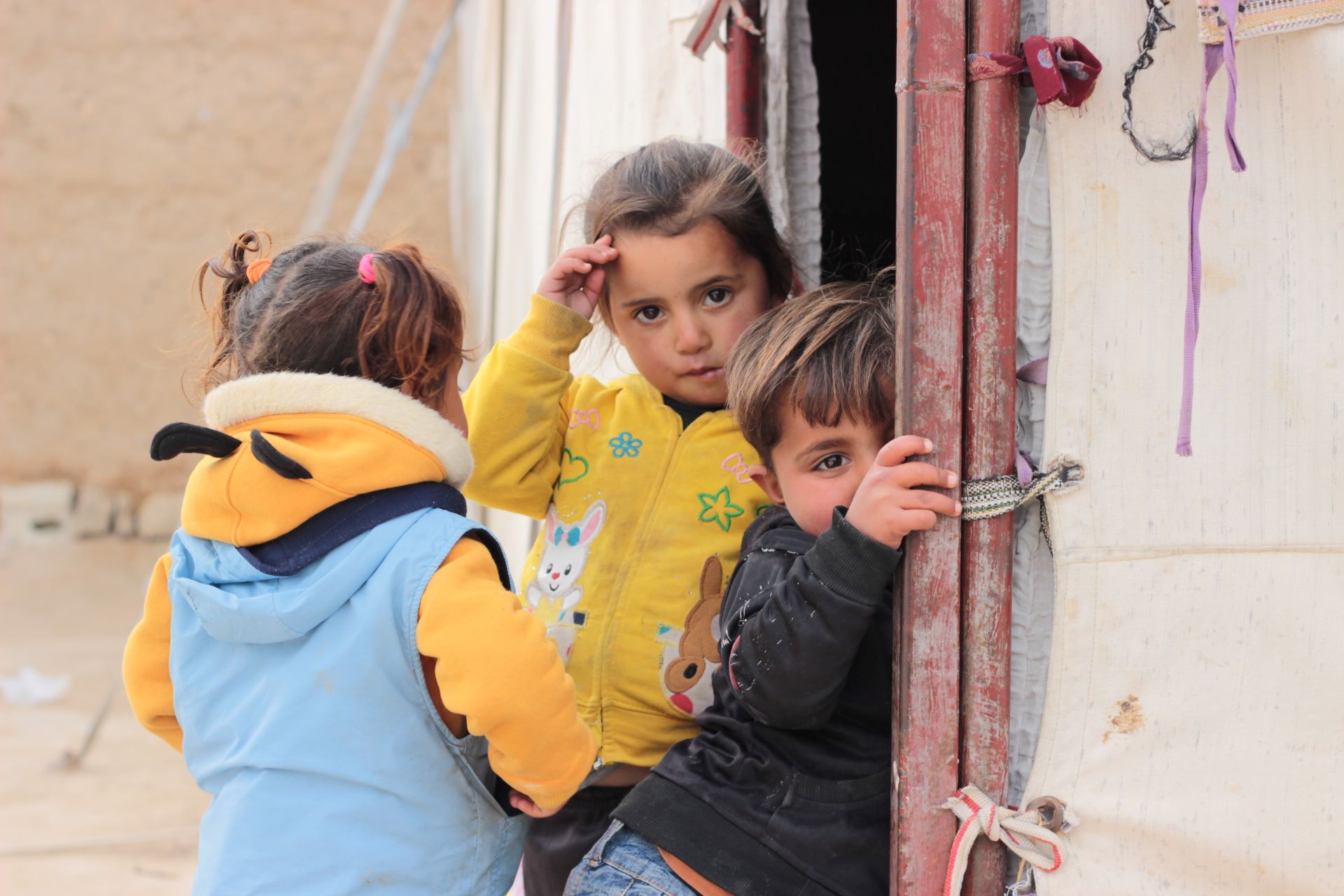 Three children (two girls, one boy) of preschool age are standing at a tent shelter. One of the girls is facing the camera, the other girl turns her back to it. The boy is facing the camera and half hides in the door frame.