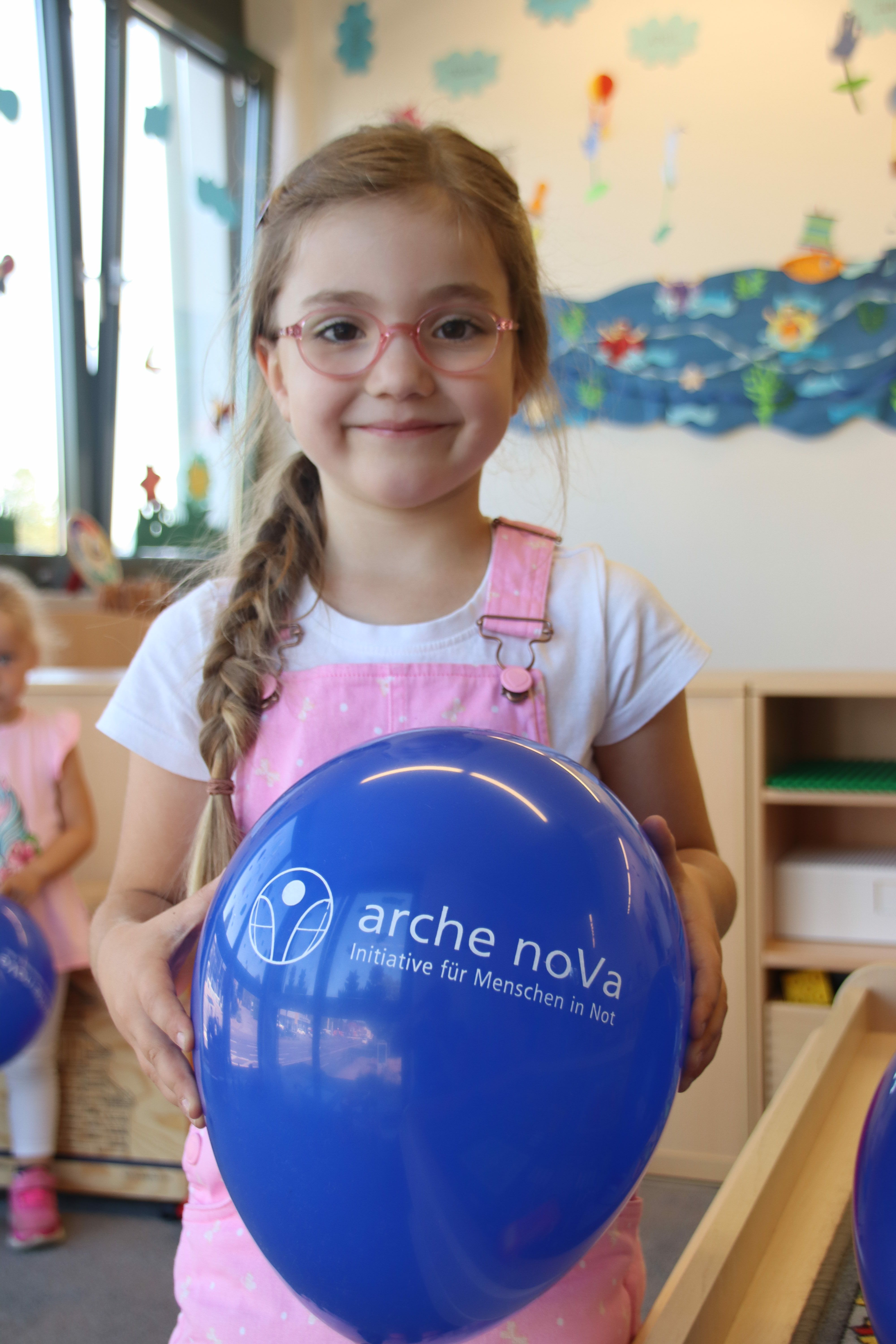 Mädchen mit Brille, geflochtenem Zopf und rosafarbener Latzhose. Das Mädchen hält einen blauen Luftballon in den Händen, der mit dem Logo von arche noVa bedruckt ist.