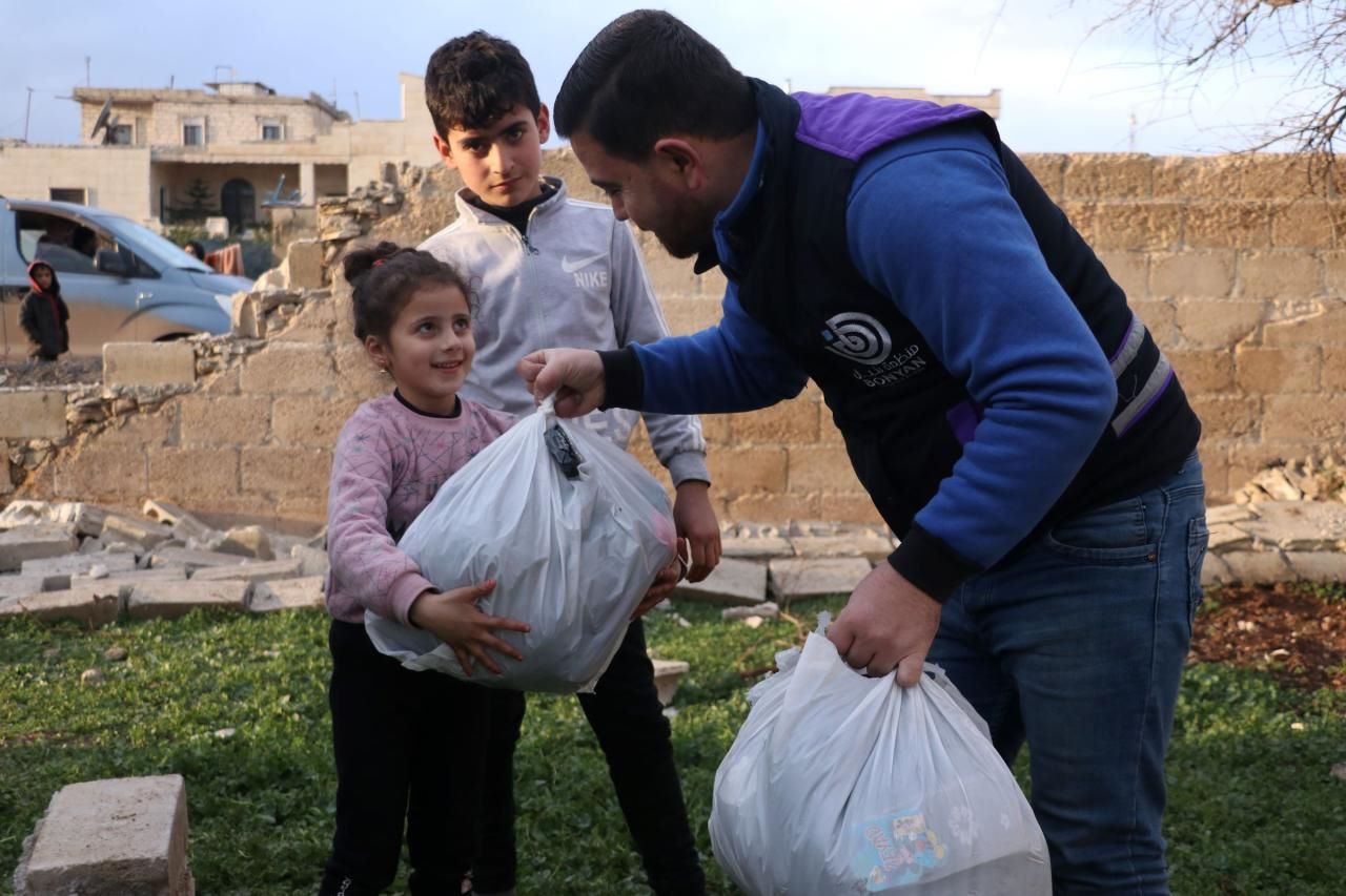 Das Bild zeigt einen Helfer unserer Partnerorganisation Takaful Al Sham. Er übergibt eine Tüte an ein Mädchen. Hinter dem Mädchen steht ein älterer Junge. Im Hintergrund sind eine zerstörte Mauer und ein Gebäude zu sehen.