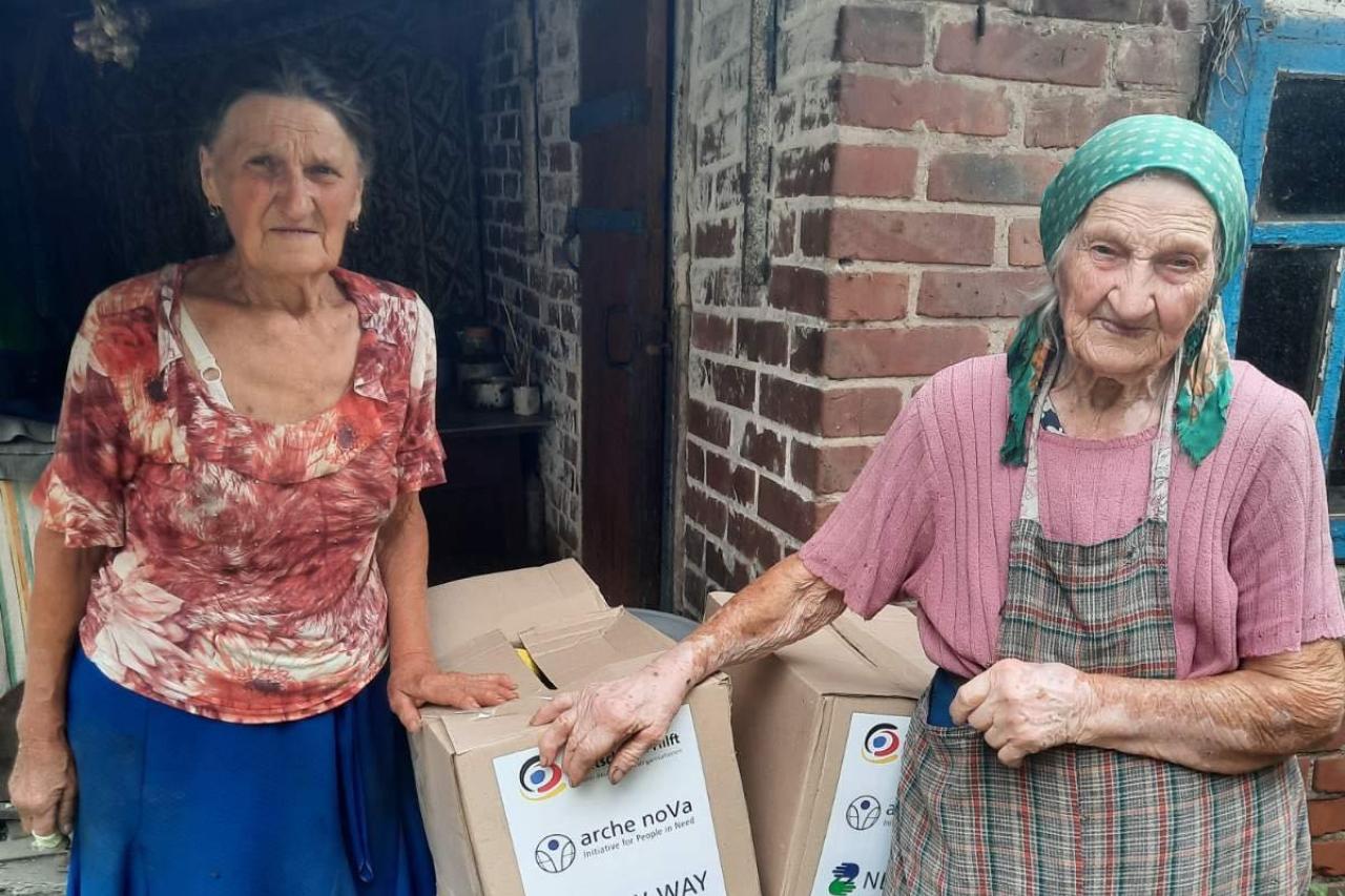 Das Bild zeigt zwei ältere Frauen, die in die Kamera blicken. Zwischen ihnen stehen zwei Kartons mit dem Logo von arche noVa, New Way und Aktion Deutschland Hilft.