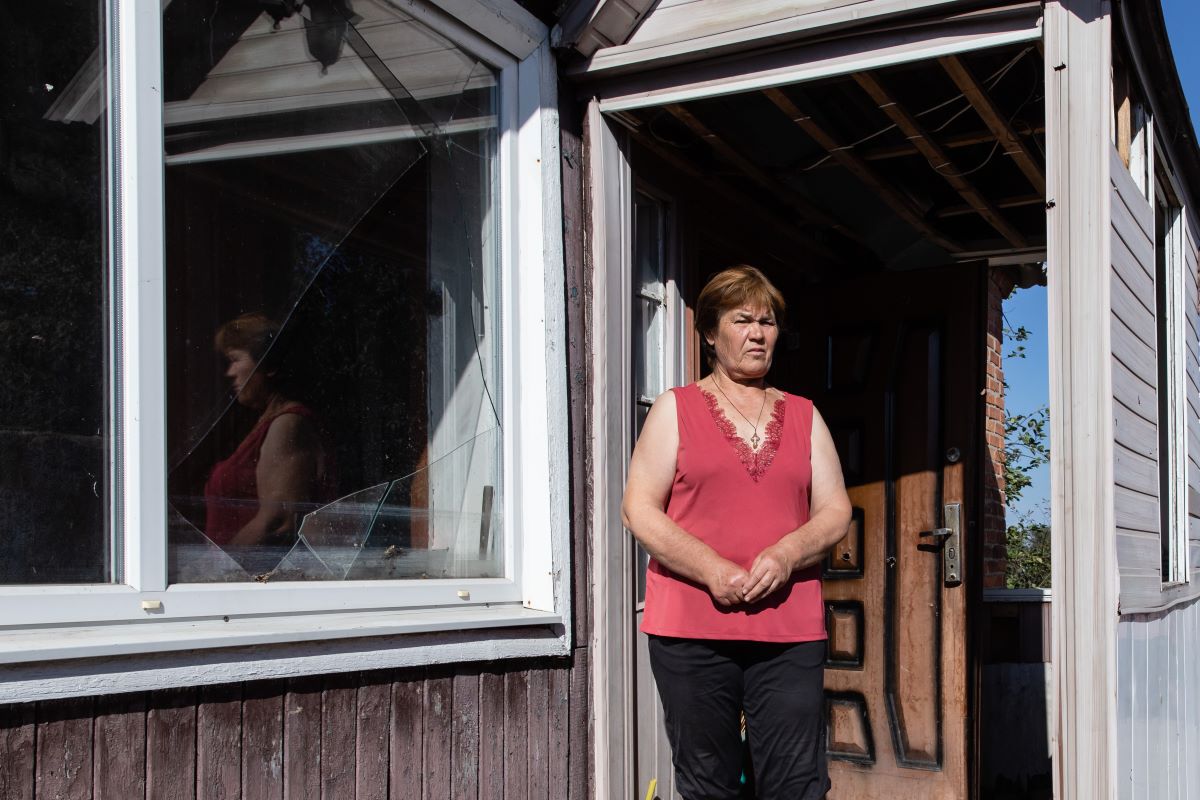Das Bild zeigt Lidiia Bohdan, eine ältere Frau aus Staryj Saltiw. Sie steht am Eingang ihres Hauses, das durch Raketen zerstört wurde. Links am Haus ist ein zerbrochenes Fenster zu sehen.