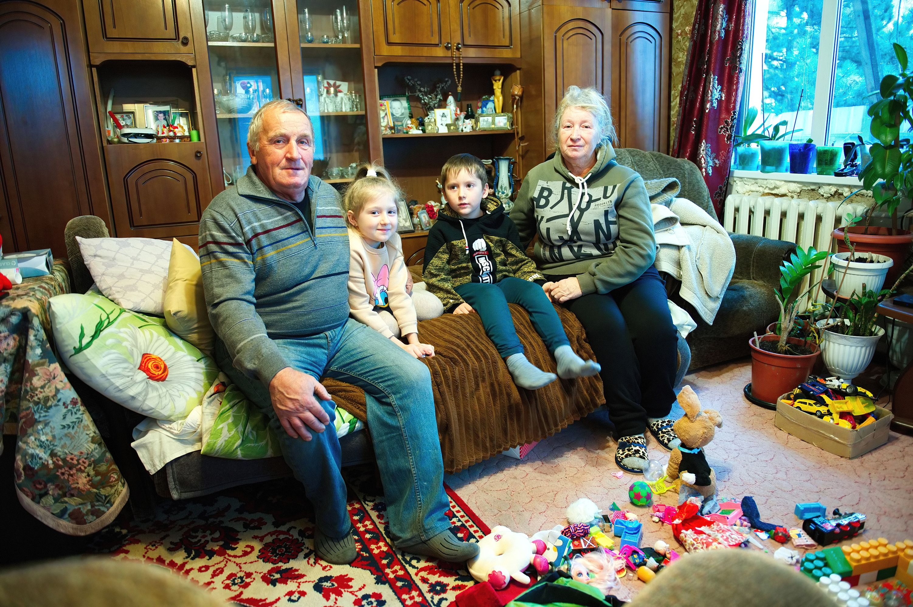 Das Bild zeigt Viktor und Lyubov Demeshko mit ihren Enkelkindern Evangelina und Mark in ihrem Wohnzimmer