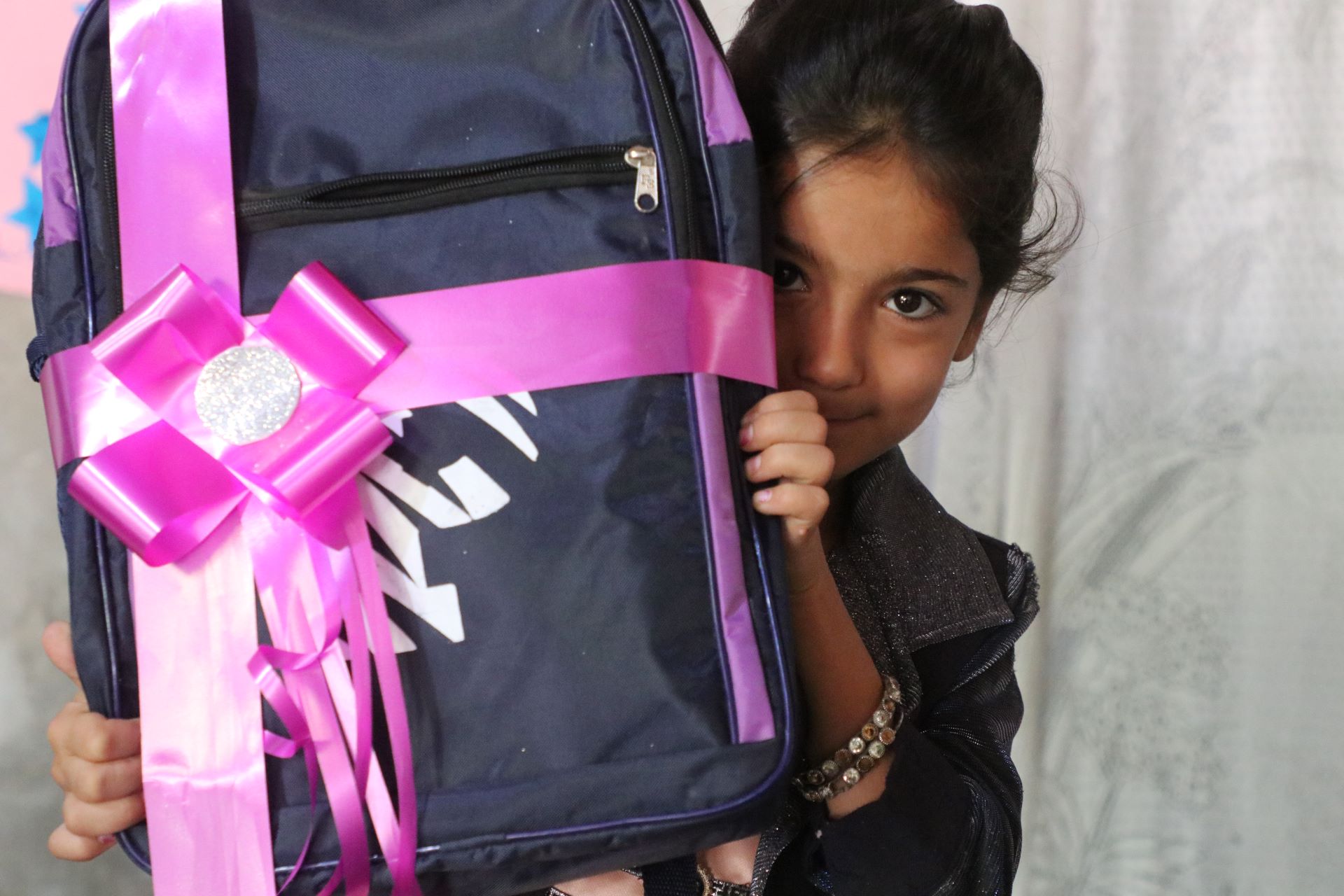 Ein Mädchen hält einen Schulrucksack mit großer pinkfarbener Schleife in die Kamera. Ihr Gesicht ist zur Hälfe hinter dem Rucksack versteckt, ihr Blick ist schüchtern.
