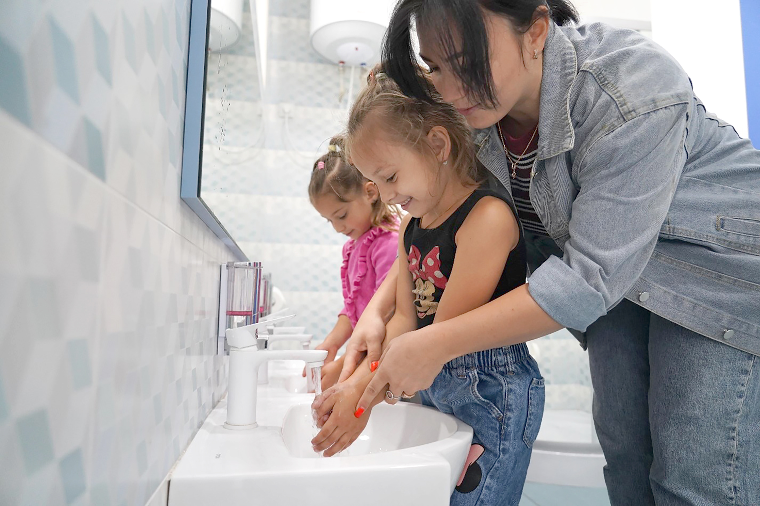 The picture shows two pre-school girls washing their hands. The girl in the foreground is being helped by a caregiver.