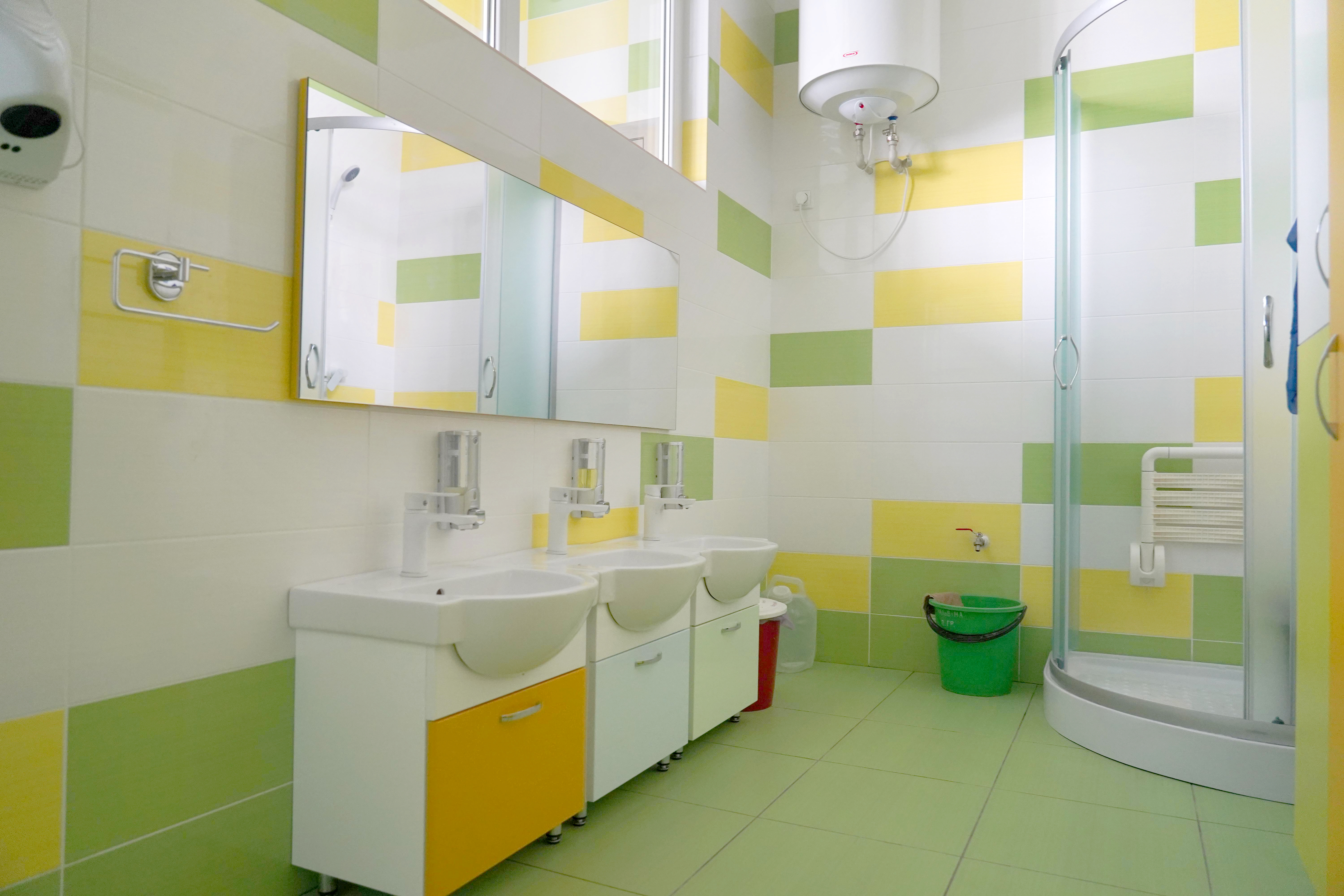 The picture shows the washroom that arche noVa has renovated. There are three washbasins on the left and a shower on the right. The fixtures, walls and floor are in light shades of green and yellow.