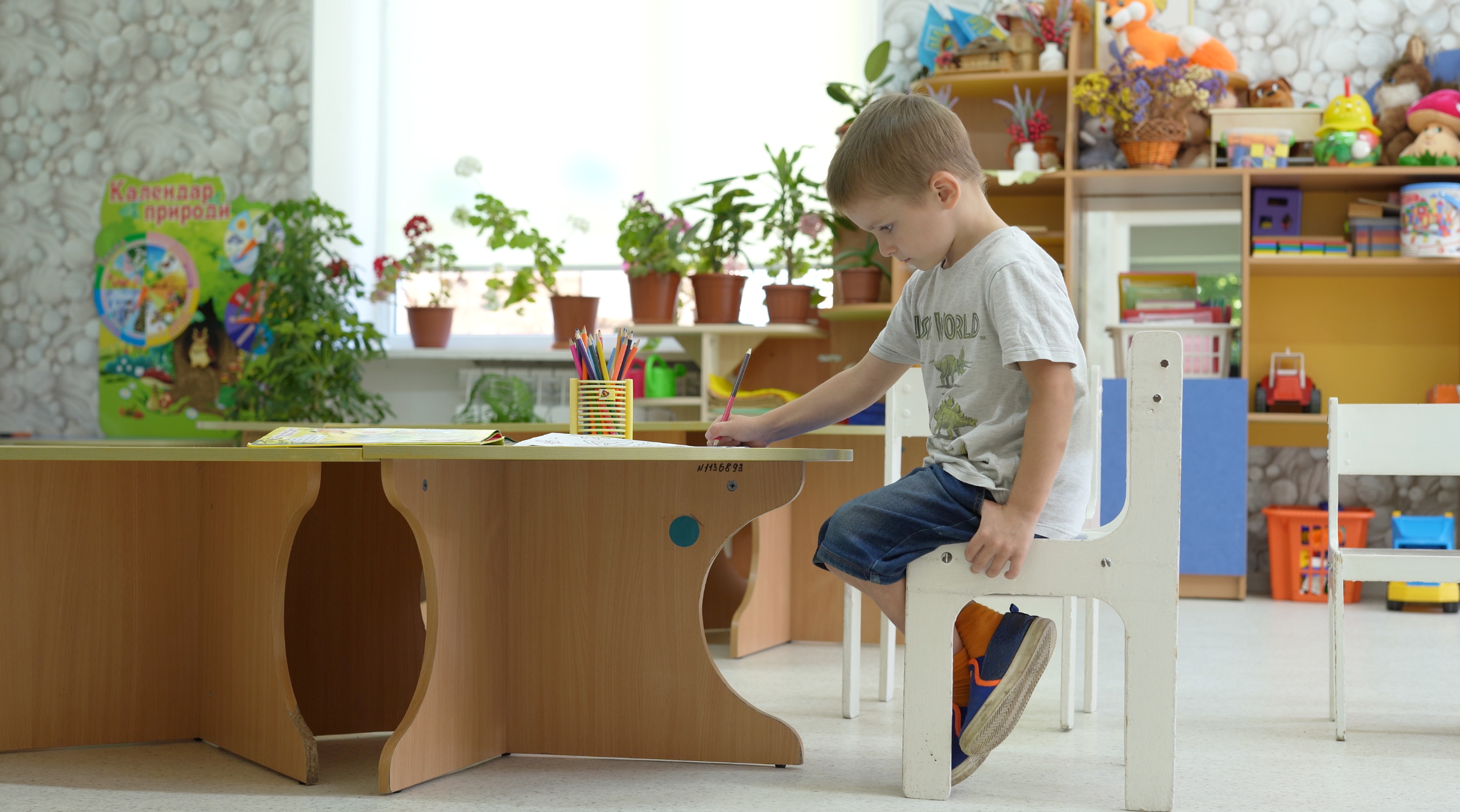 Innenraum der Kita. Kyrylo sitzt auf einem kleinen weißen Stuhl vor einem Kindertisch. Er schaut auf das Blatt auf dem Tisch, auf dem er mit der rechten Hand schreibt. Im Hintergrund sind Schränke, ein Fenster und Zimmerpflanzen