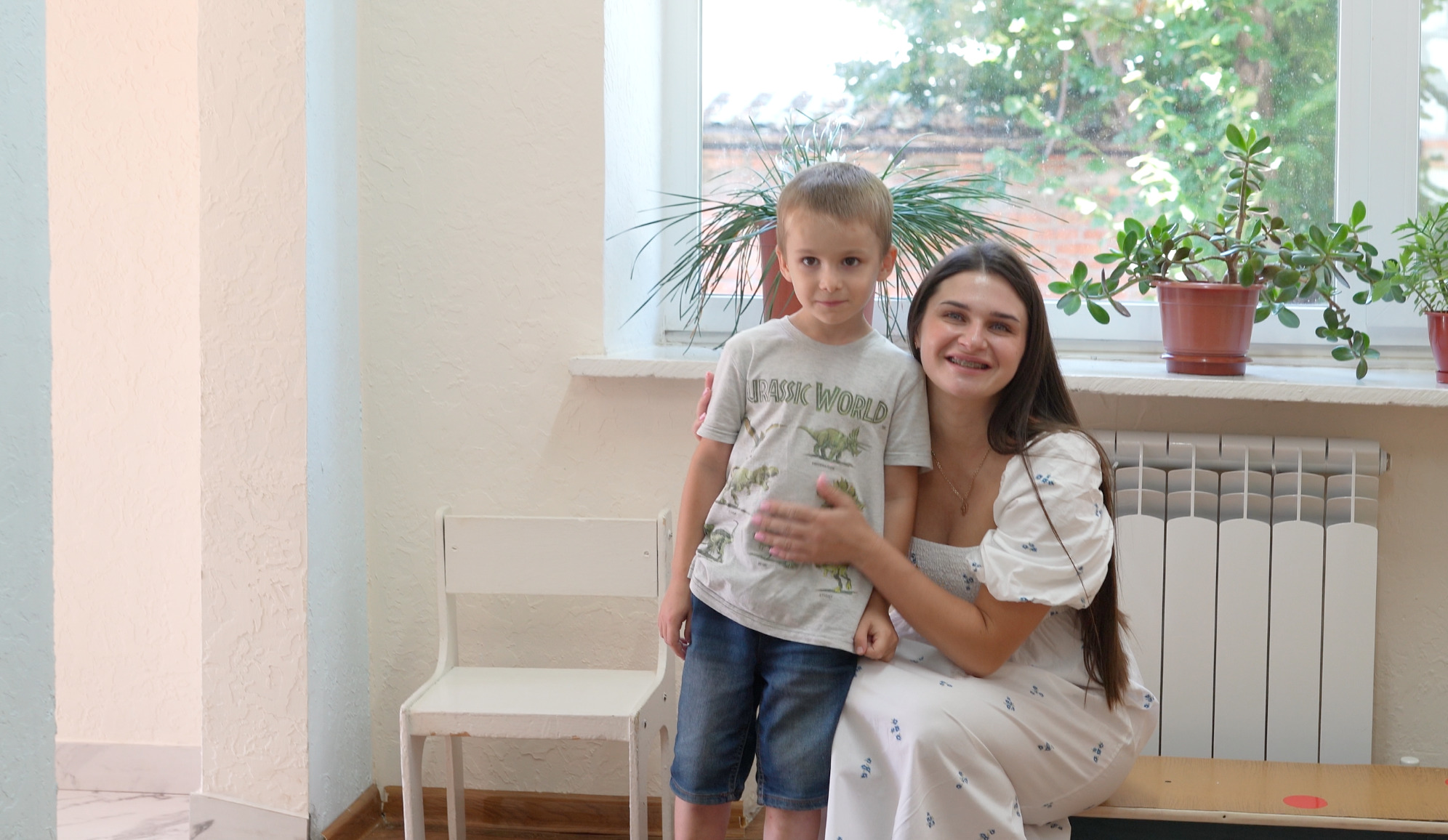 Porträt von Kyrylo und Yuliia. Kyrylo steht in der Mitte des Bildes, Yuliia sitzt rechts und umarmt ihn. Im Hintergrund sind ein weißer Stuhl, ein Fenster und Zimmerpflanzen