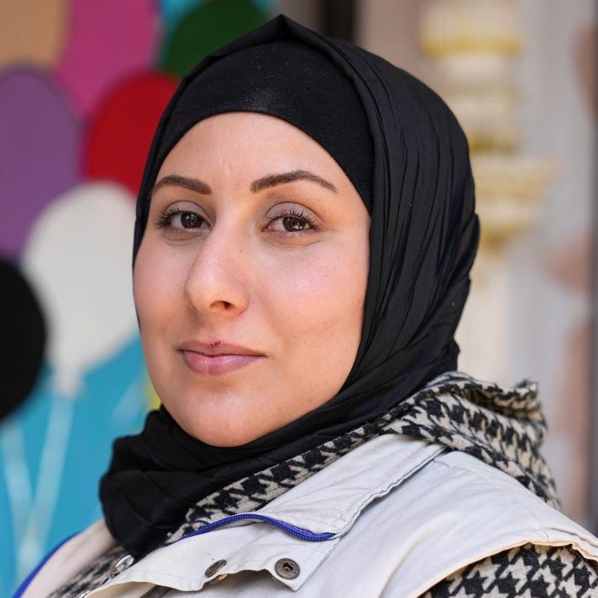 The picture shows a portrait of the Bonyan helper Sana. She is wearing a black headscarf, a beige-coloured waistcoat and black and white patterned outerwear.