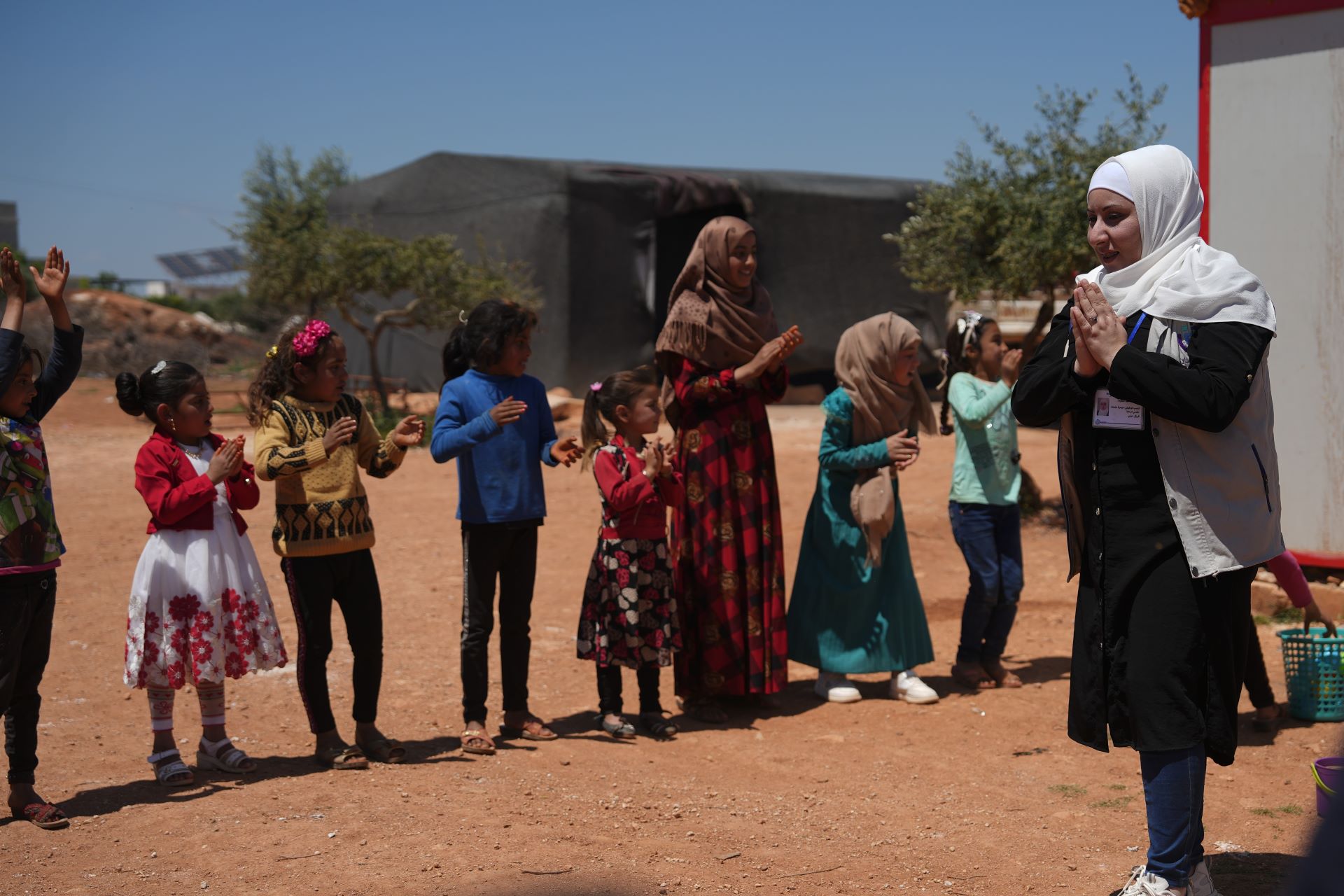 Das Bild zeigt die Kinderschutzbeauftragte Sana Abdulatif mit einer Gruppe von Kindern. Sie stehen draußen, alle klatschen in die Hände.