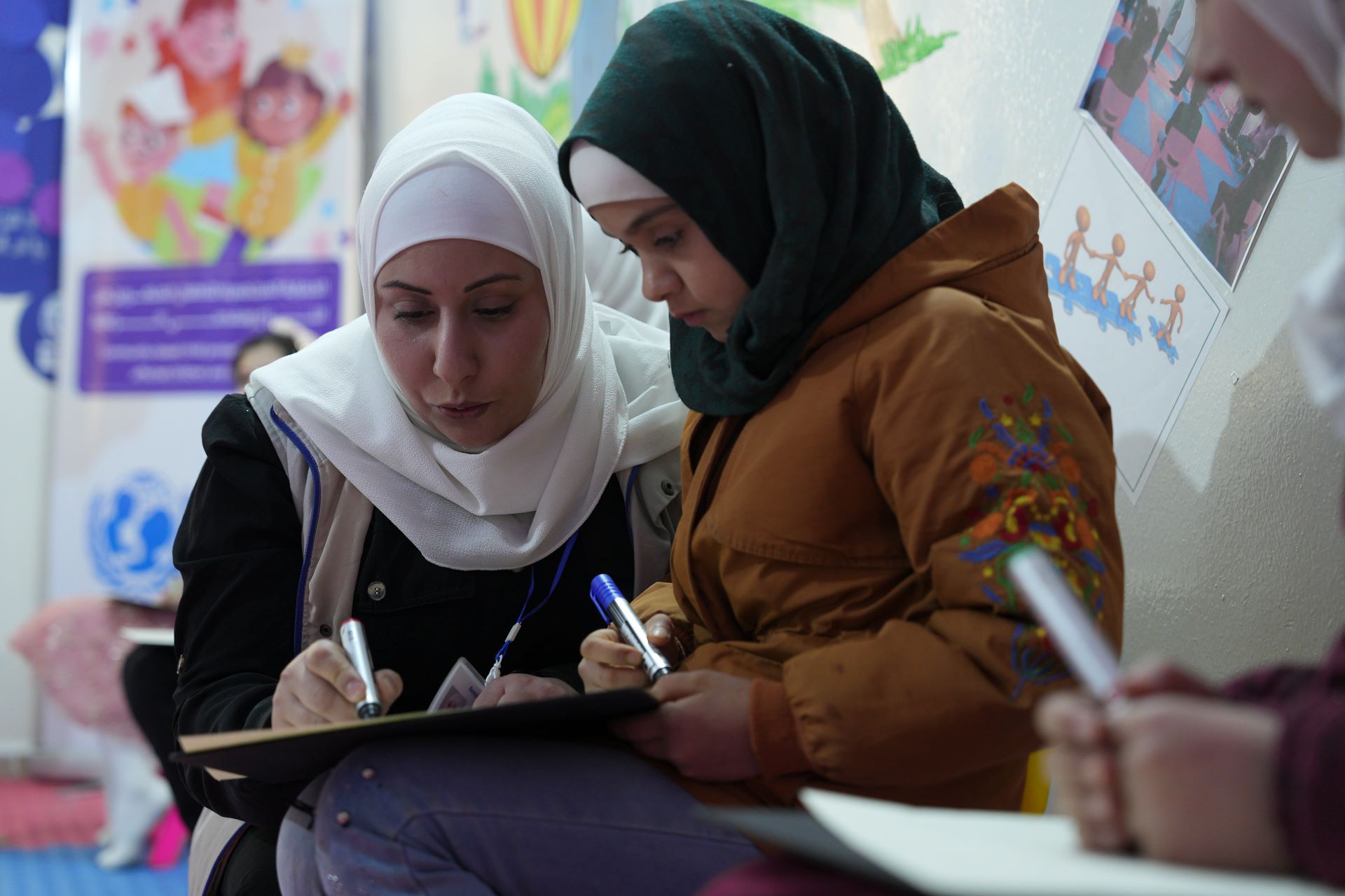 Das Bild zeigt die Kinderschutzbeauftragte Sana Abdulatif neben einem Mädchen. Beide sitzen und halten einen Stift in der Hand. Sana schreibt auf ein Blatt Papier, sie scheint dem Mädchen etwas zu erklären.