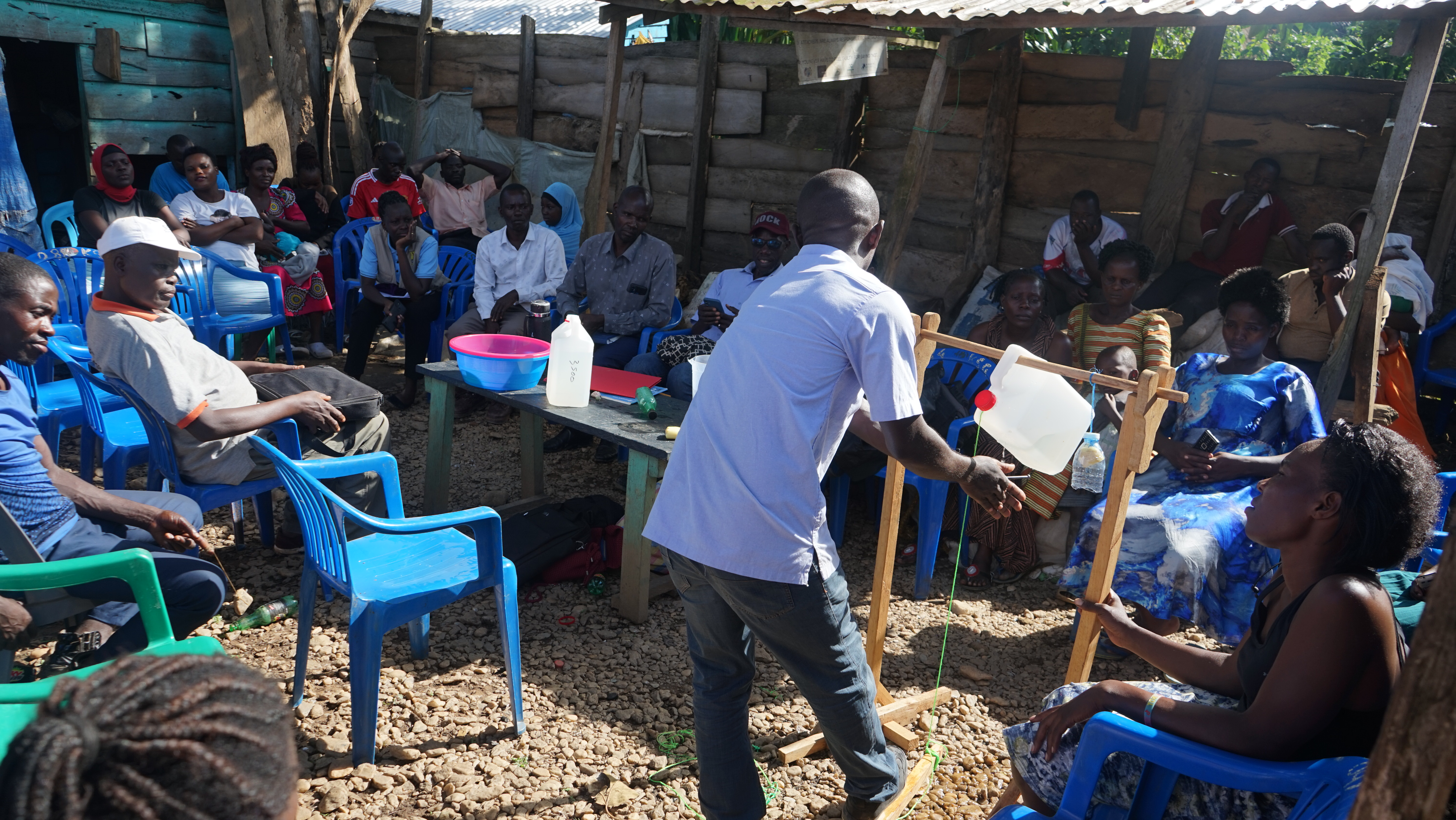 Eine kleine Gruppe Menschen sitzt auf Stühlen im Kreis. Sie gucken in die Mitte, wo ein Mann seine Hände wäscht, wobei das Wasser aus einem Kanister fließt.