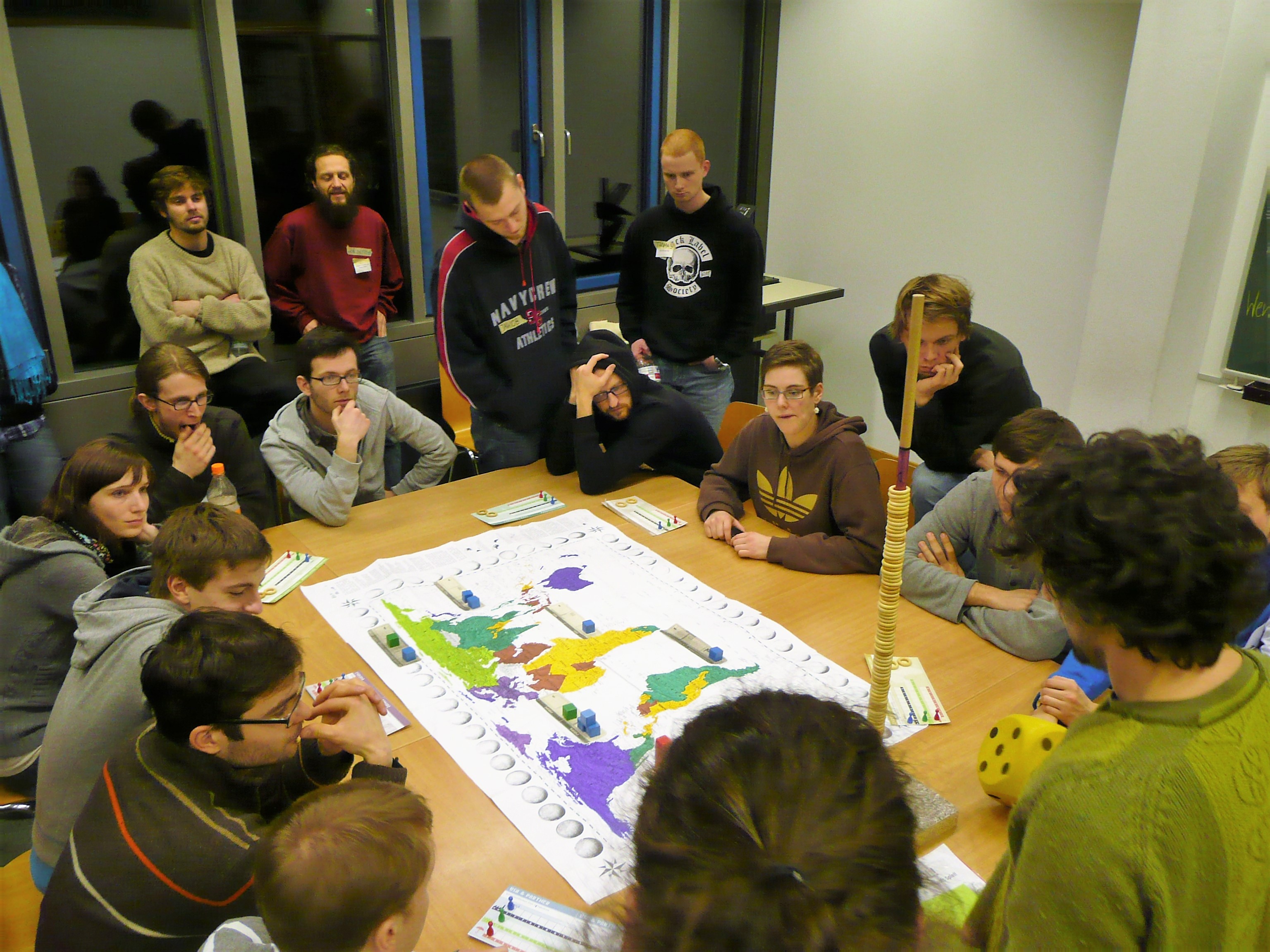 Studierende sind um einen Tisch versammelt, auf dem eine Weltkarte liegt. Ein großer Würfel und Spielfiguren sind zu sehen.