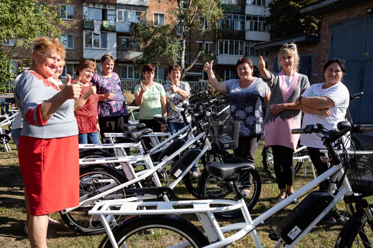 Das Bild zeigt die Mitarbeiterinnen des Sozialdienstes in Ochtyrka. Die Frauen stehen neben mehreren Elektrofahrrädern. Einige von ihnen halten den Daumen nach oben.