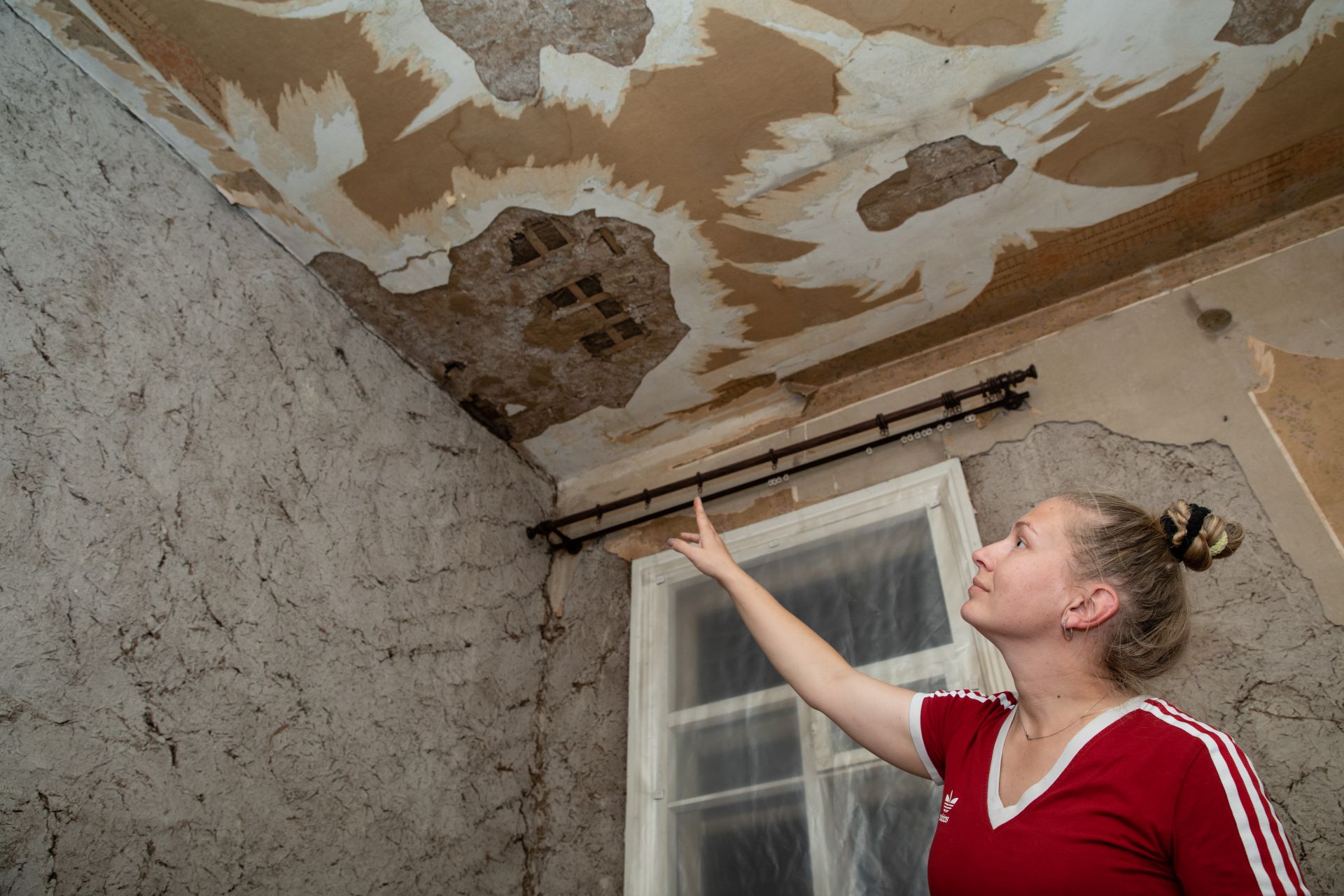 Nadiia Novytska zeigt auf eine undichte Stelle in der Decke ihres Hauses. Der Putz ist beschädigt, das Fenster im Hintergrund mit Folie abgeklebt.