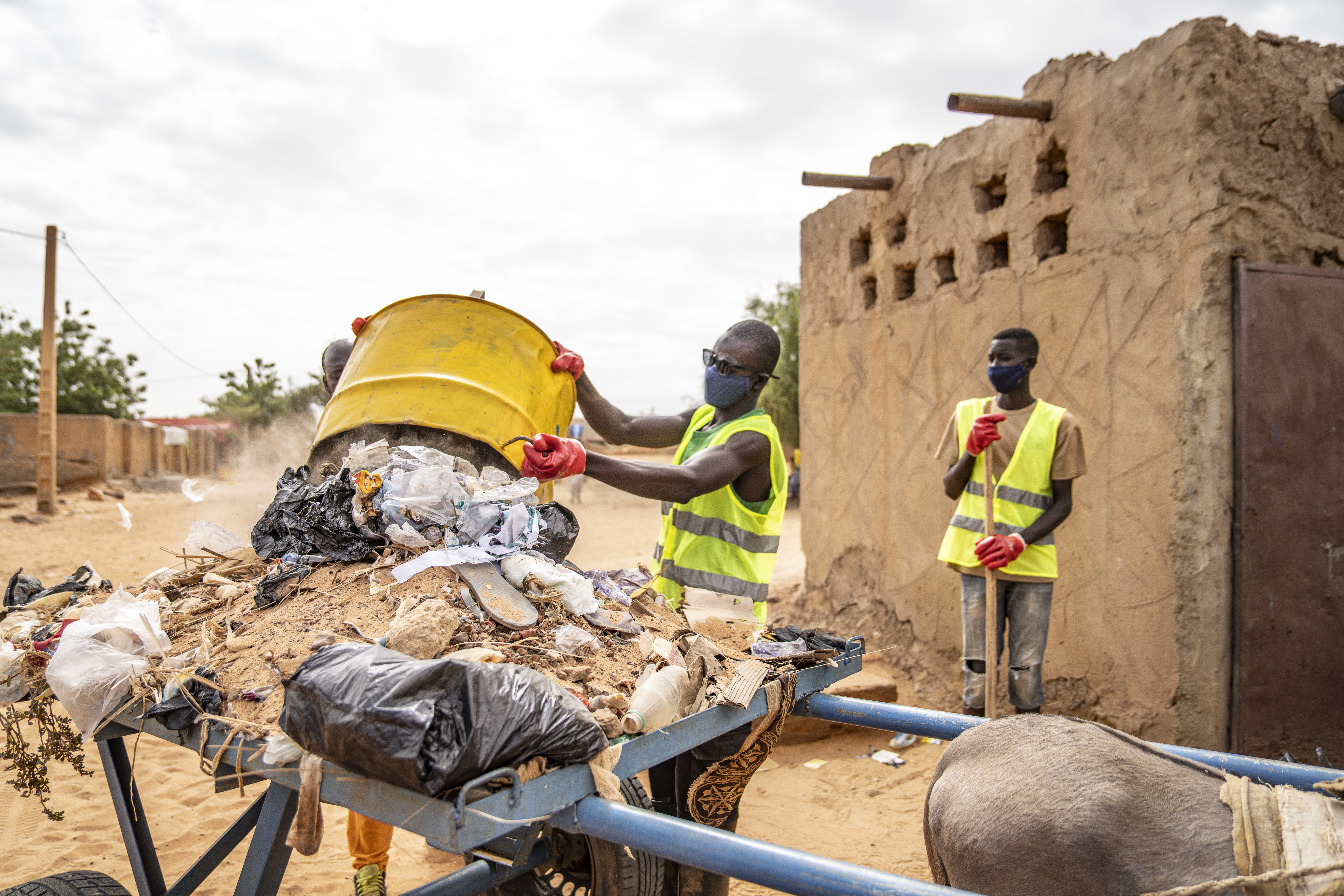 Das Foto zeigt zwei Männer in Warnwesten und mit Schutzmasken. Einer von ihnen belädt einen Karren mit Abfall, der andere steht im Hintergrund und hält einen Besen in der Hand.