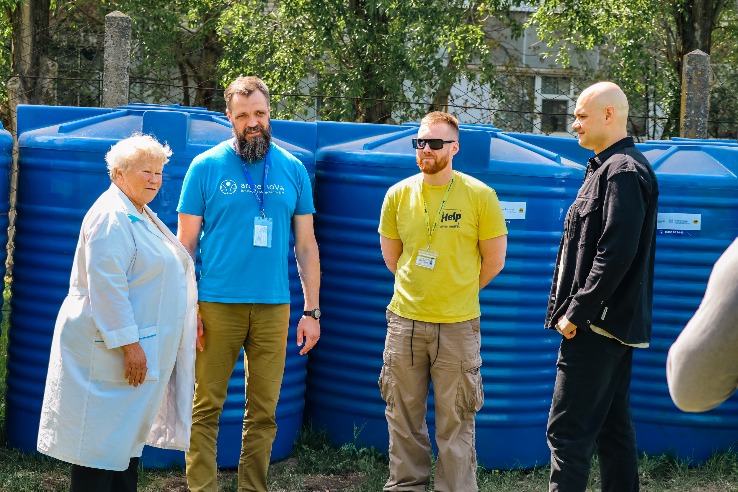 Das Bild zeigt eine Mitarbeiterin des örtlichen Wasserversorgers von Selenodolsk und Dmytro Drizhd, Leiter des Büros von arche noVa in der Ukraine, einen Mitarbeitenden von Help – Hilfe zur Selbsthilfe sowie den Bürgermeister Dmytro Nevesely. Alle stehen vor einer Reihe von Wassertanks. 