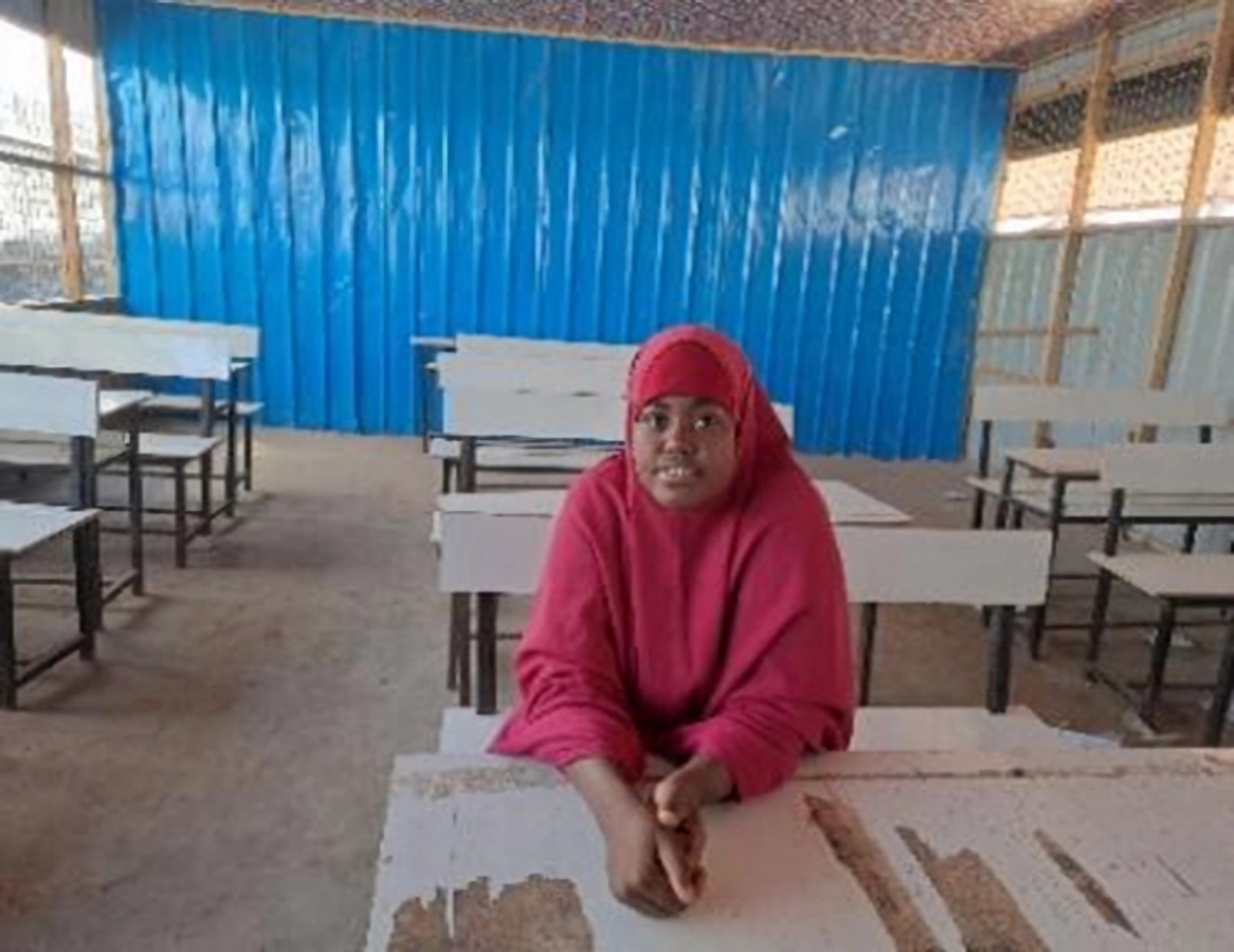 Girl in red top and headscarf sitting at a table, looking at the camera, in the background more school desks and a blue wall