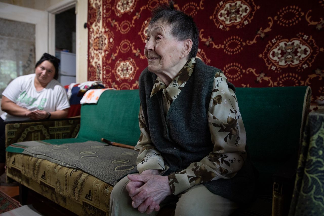The picture shows Valentyna Mykolaivna sitting on her couch. She holds her hands in her lap. In the background you can see Alina Kovalenko, who visits her regularly.