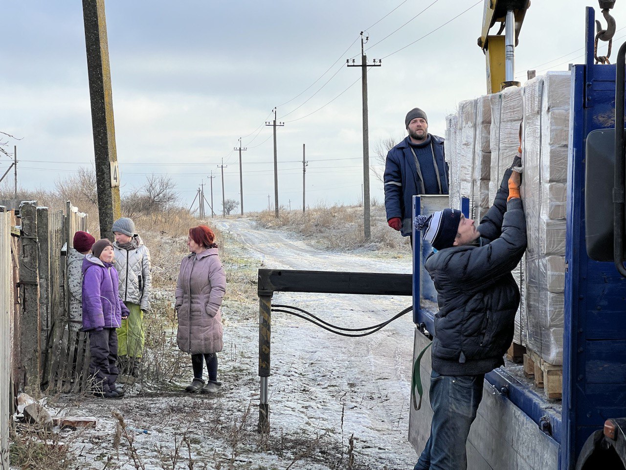 Das Bild zeigt zwei Männer an einen Transporter, der mit Holzbriketts beladen ist. Am Boden liegt etwas Schnee, im Hintergrund sind eine Frau und drei Kinder zu sehen.