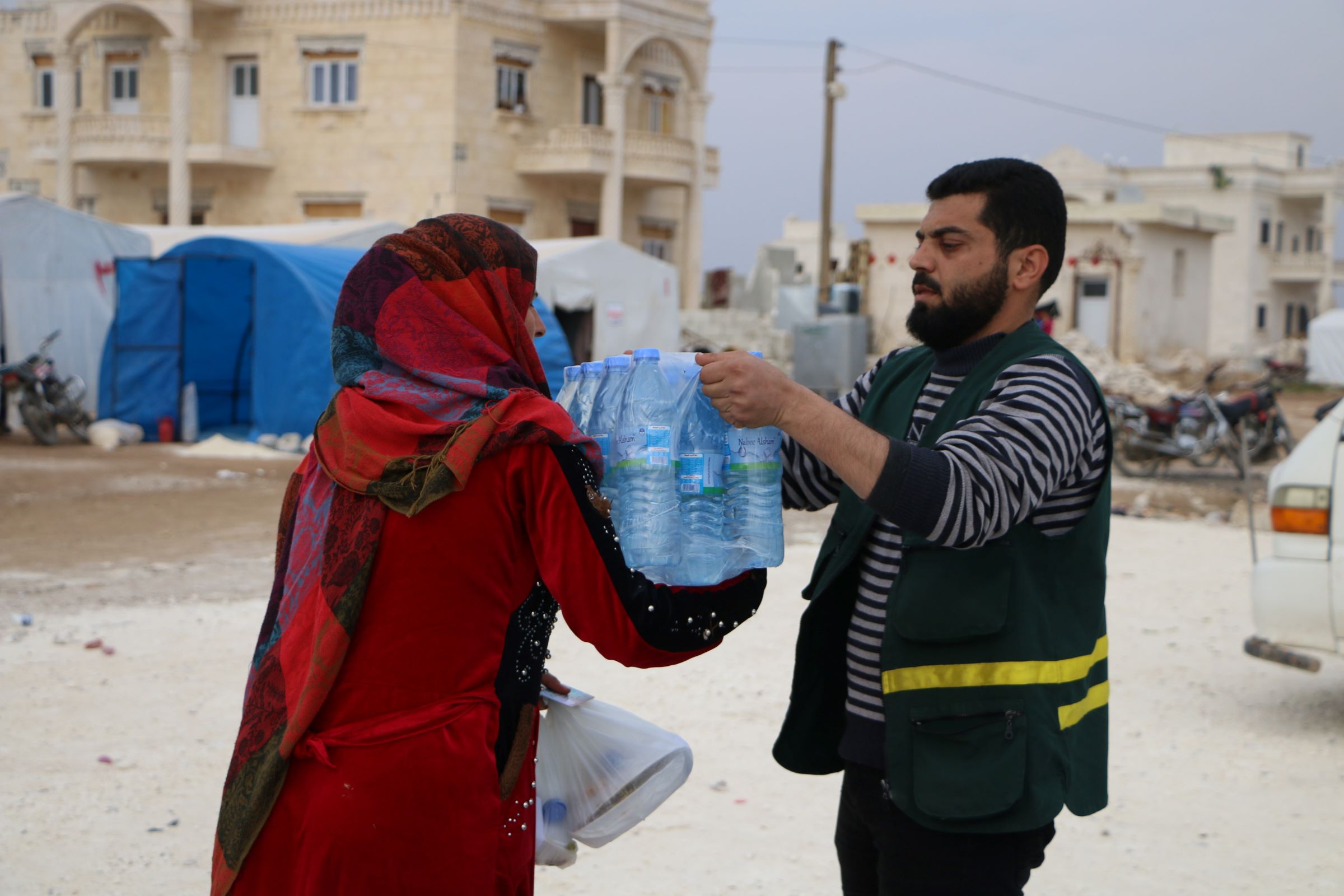An emergency worker from our local partner organization Takaful Al-Sham hands a six-pack of water bottles to a woman. Tent shelters can be seen in the background.