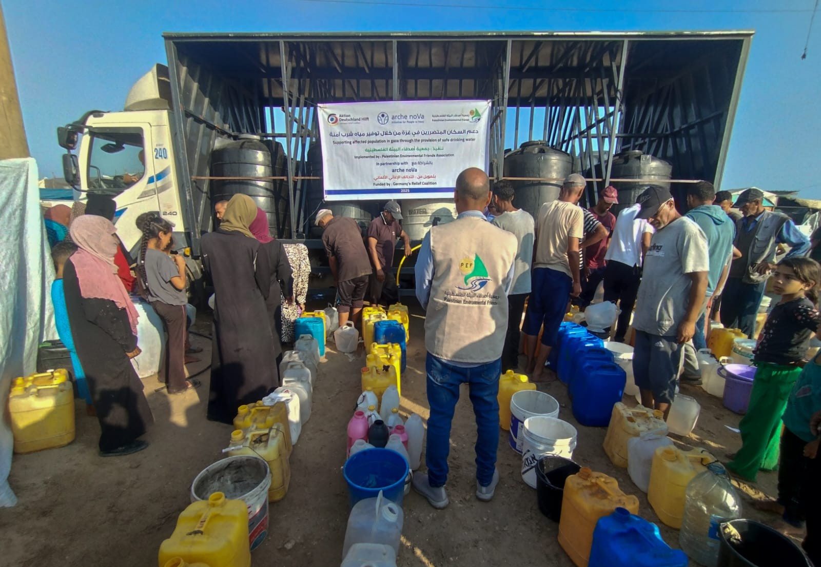 Das Bild zeigt Wasserkanister in unterschiedlichen Farben, die in einer Reihe aufgestellt sind. Um sie herum viele Menschen, im Hintergrund ist ein Lkw mit Wassertanks zu sehen.