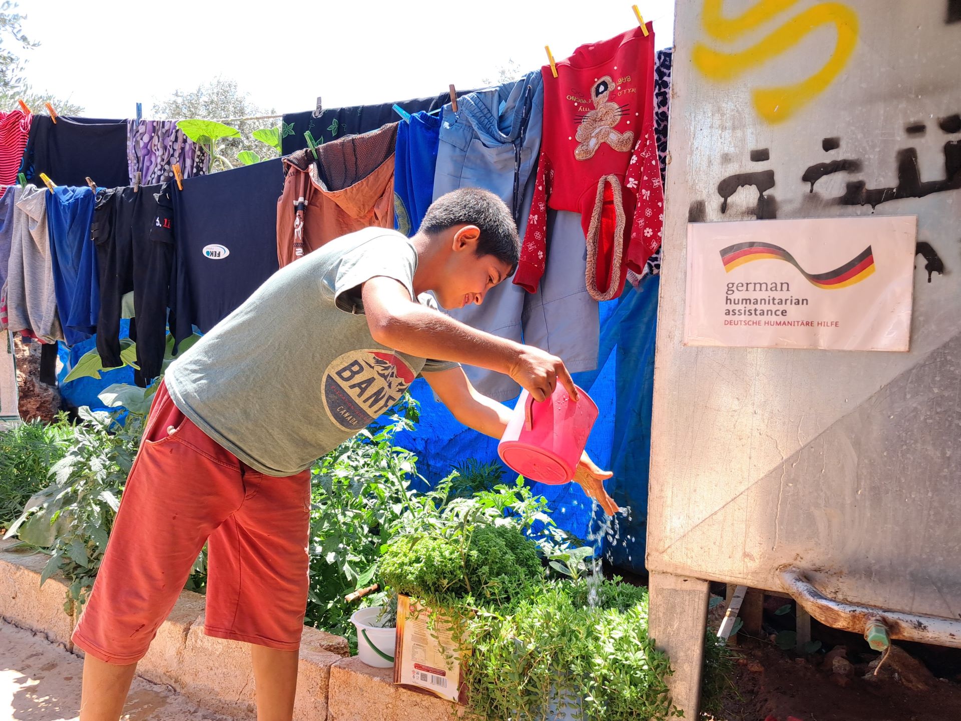 Das Bild zeigt einen Jungen, der verschiedene Pflanzen gießt. Neben ihm steht ein Wassertank aus Metall, im Hintergrund ist Wäsche zu sehen.