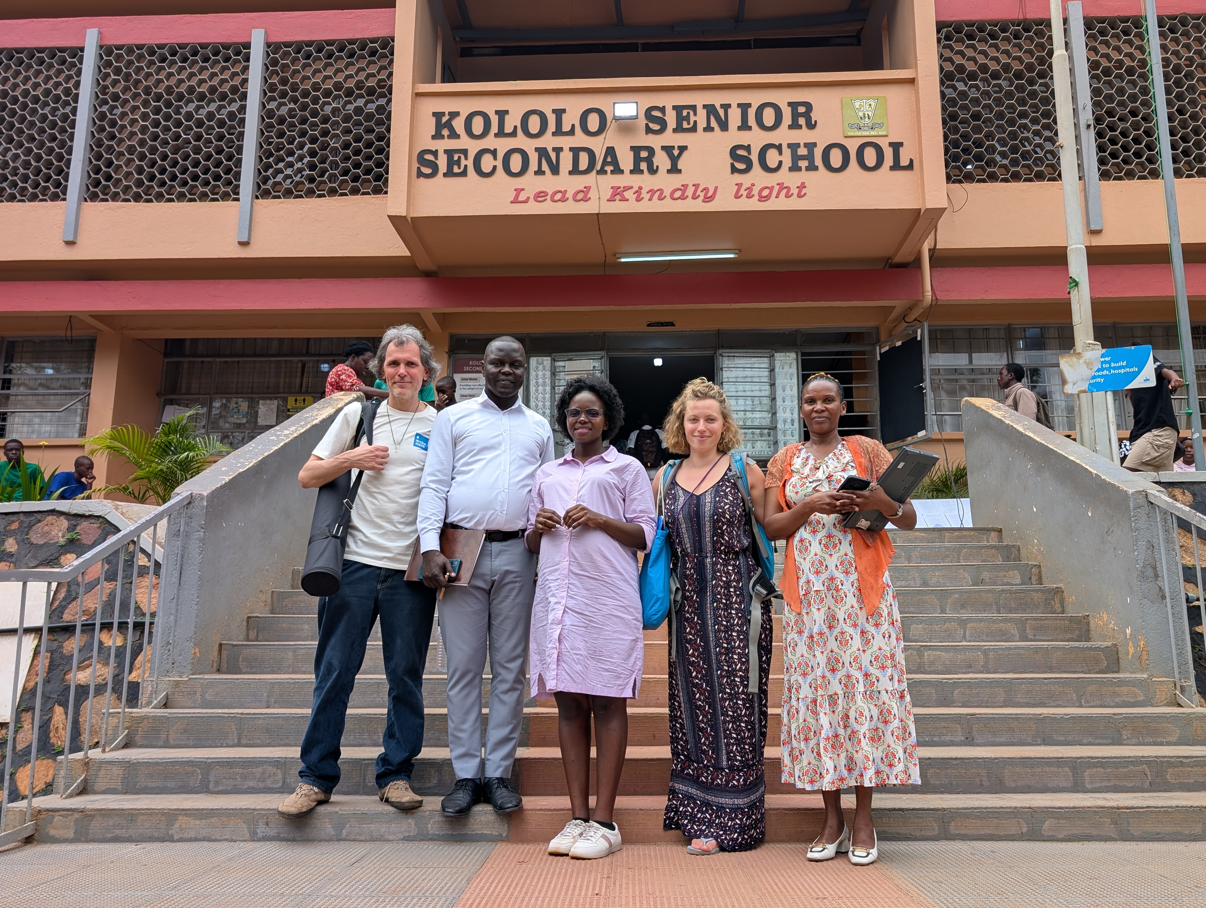 Eie Gruppe von Personen posiert für ein Gruppenfoto. Im Hintergrund ist eine Treppe zu sehen, die zum Schulgebäude der Kololo Senior Secondary School führt.