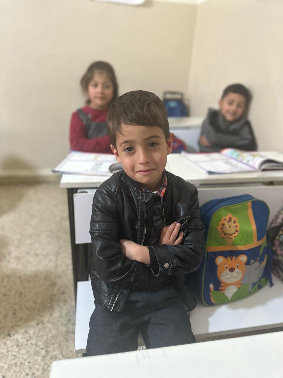 Ein ungefähr 6 Jahre alter Junge sitzt auf einer Schulbank im Klassenzimmer. Er hat die Arme verschränkt und lächelt
