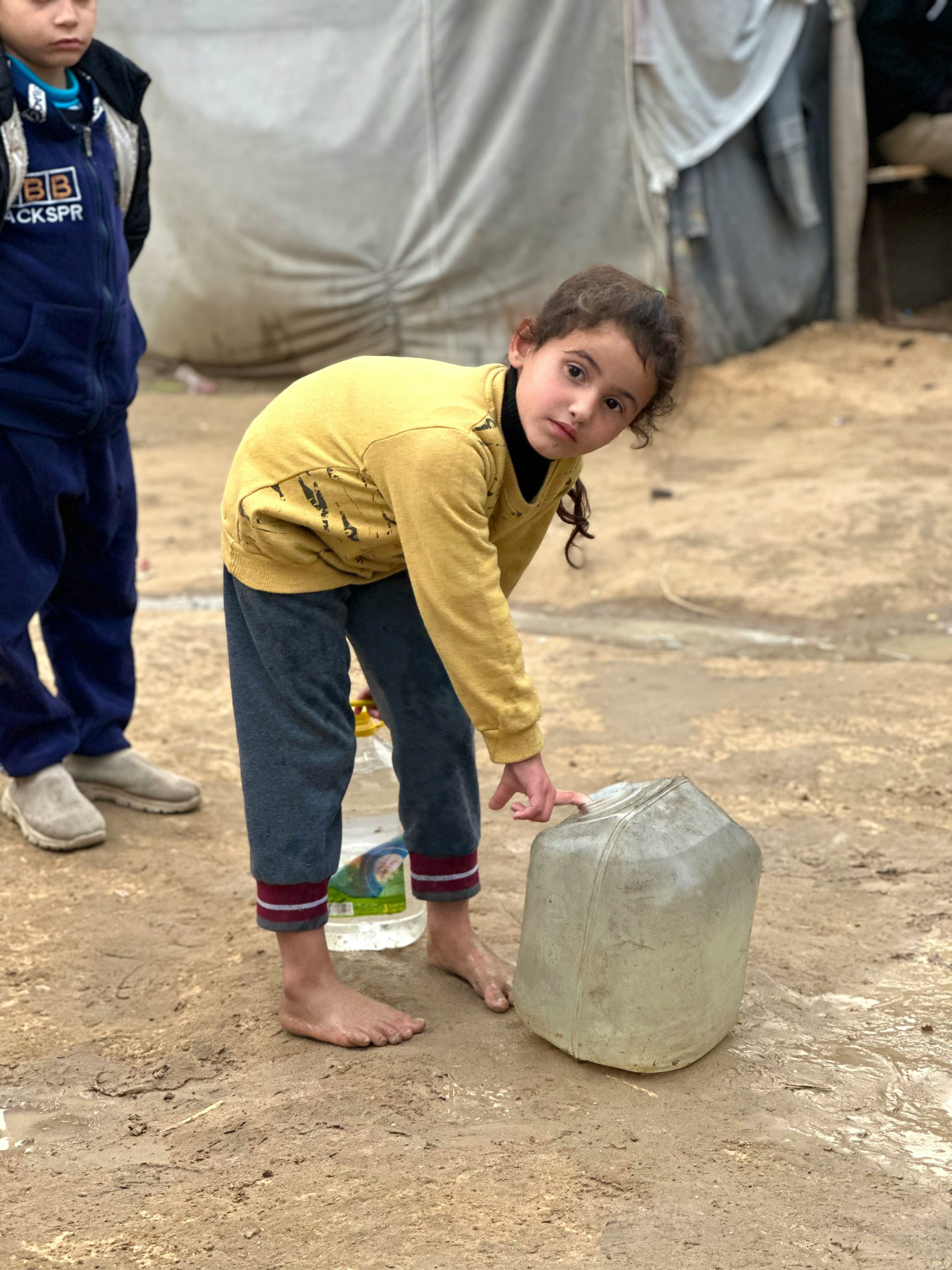 Mädchen im Grundschulalter in leicht gebeugter Haltung neben einem am Boden stehenden Wasserkanister, den sie mit dem Finger antippt. Sie blickt direkt in die Kamera.
