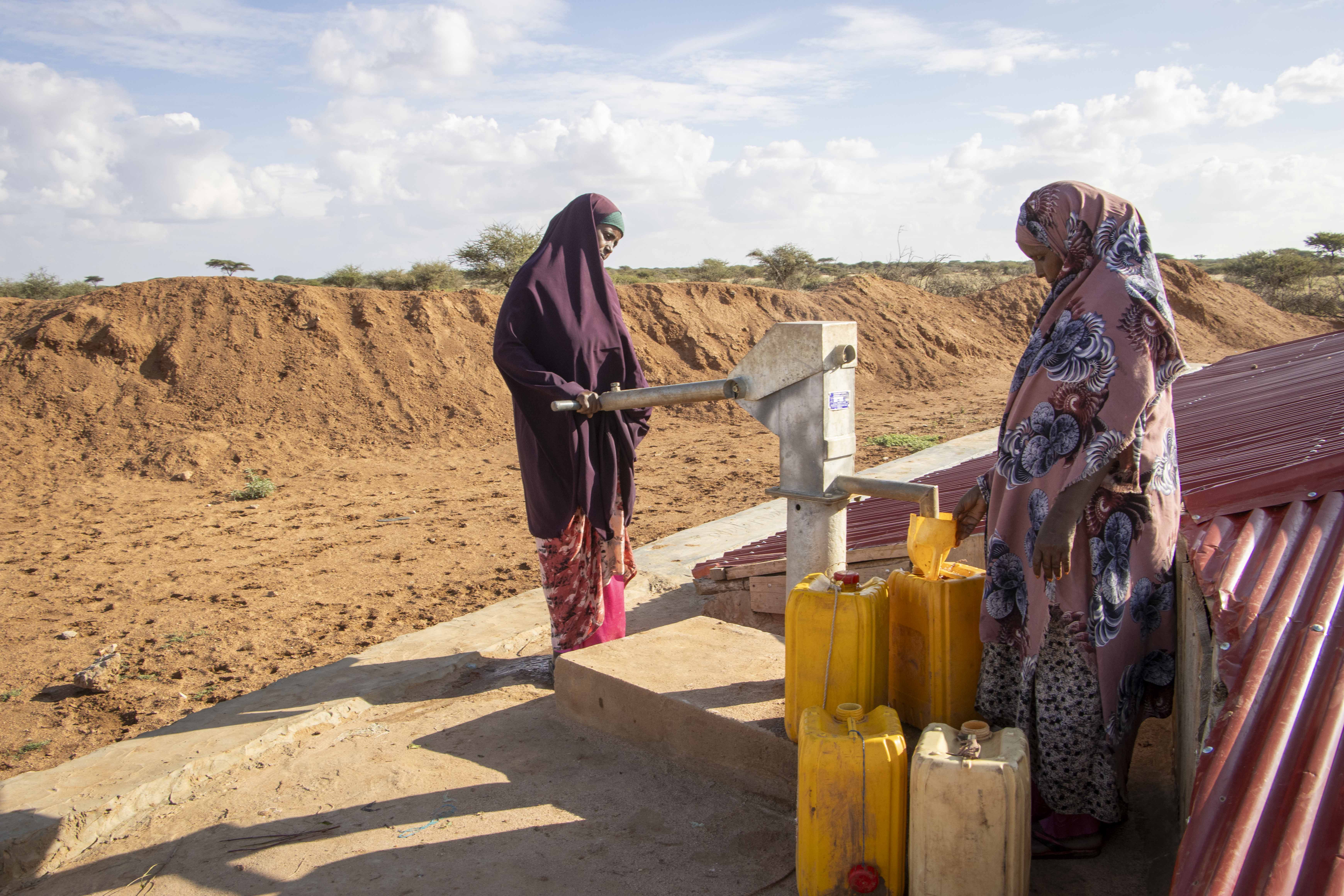 Frauen an einer Handpumpe, rechts am Bildrand ein Birka Wasserspeicher