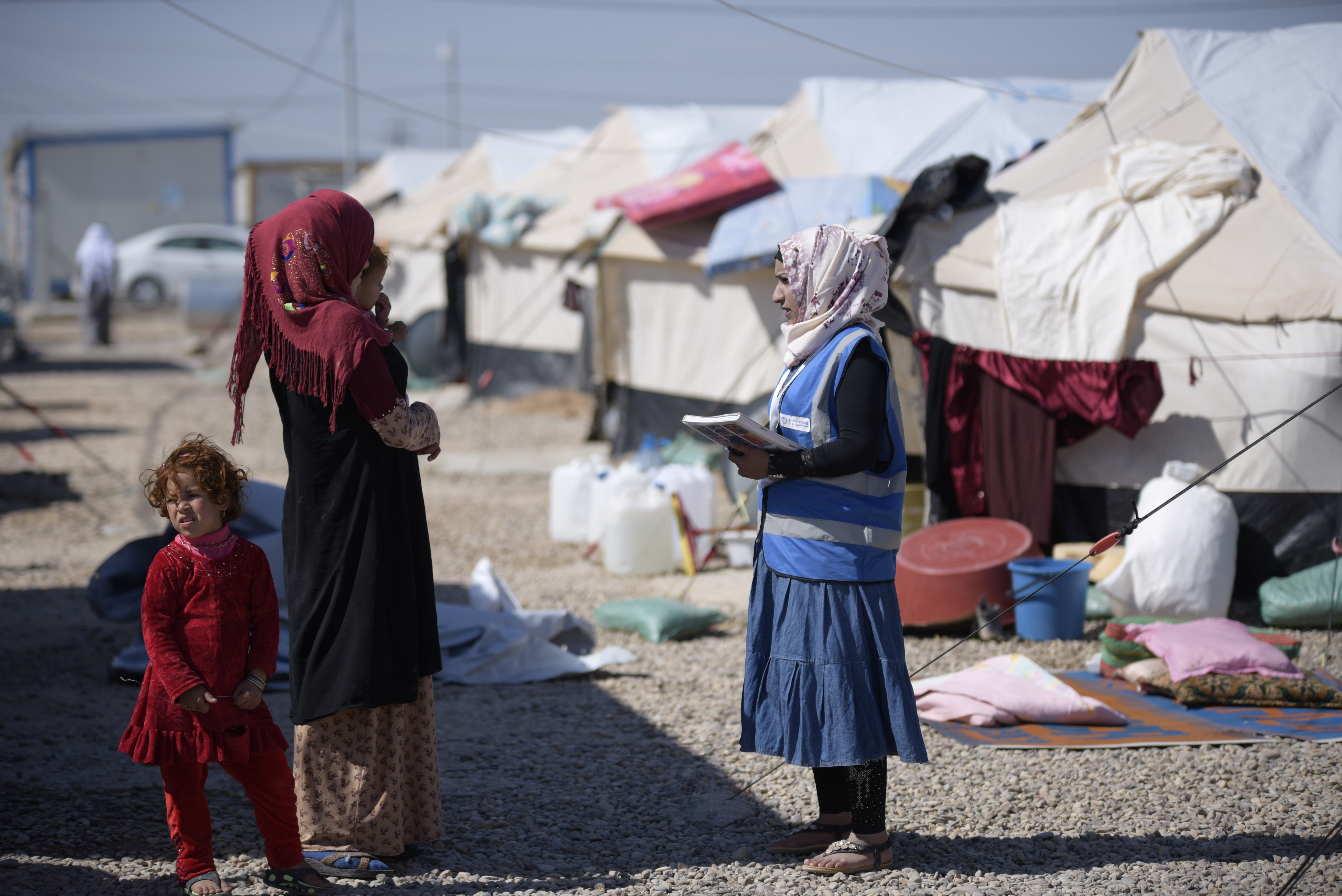 Eine Hygienepromoterin im Gespräch mit einer Mutter, die zwei Kleinkinder bei sich hat, im Hintergrund Zelte eines Camps