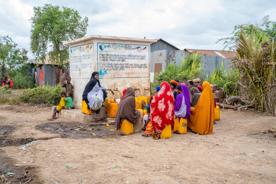 Eine Gruppe von Frauen sitzt auf gelben Kanistern an einer Wasserausgabestelle. Aus Wasserhähnen läuft Wasser in weitere gelbe Kanister.