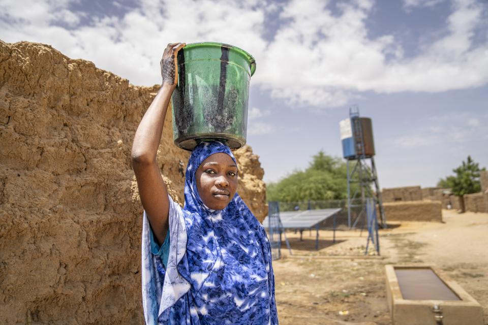 Das Foto zeigt eine junge Frau. Sie trägt einen Eimer auf ihrem Kopf und blickt selbstbewusst in die Kamera. Im Hintergrund ist ein Wasserversorgungssystem zu sehen.